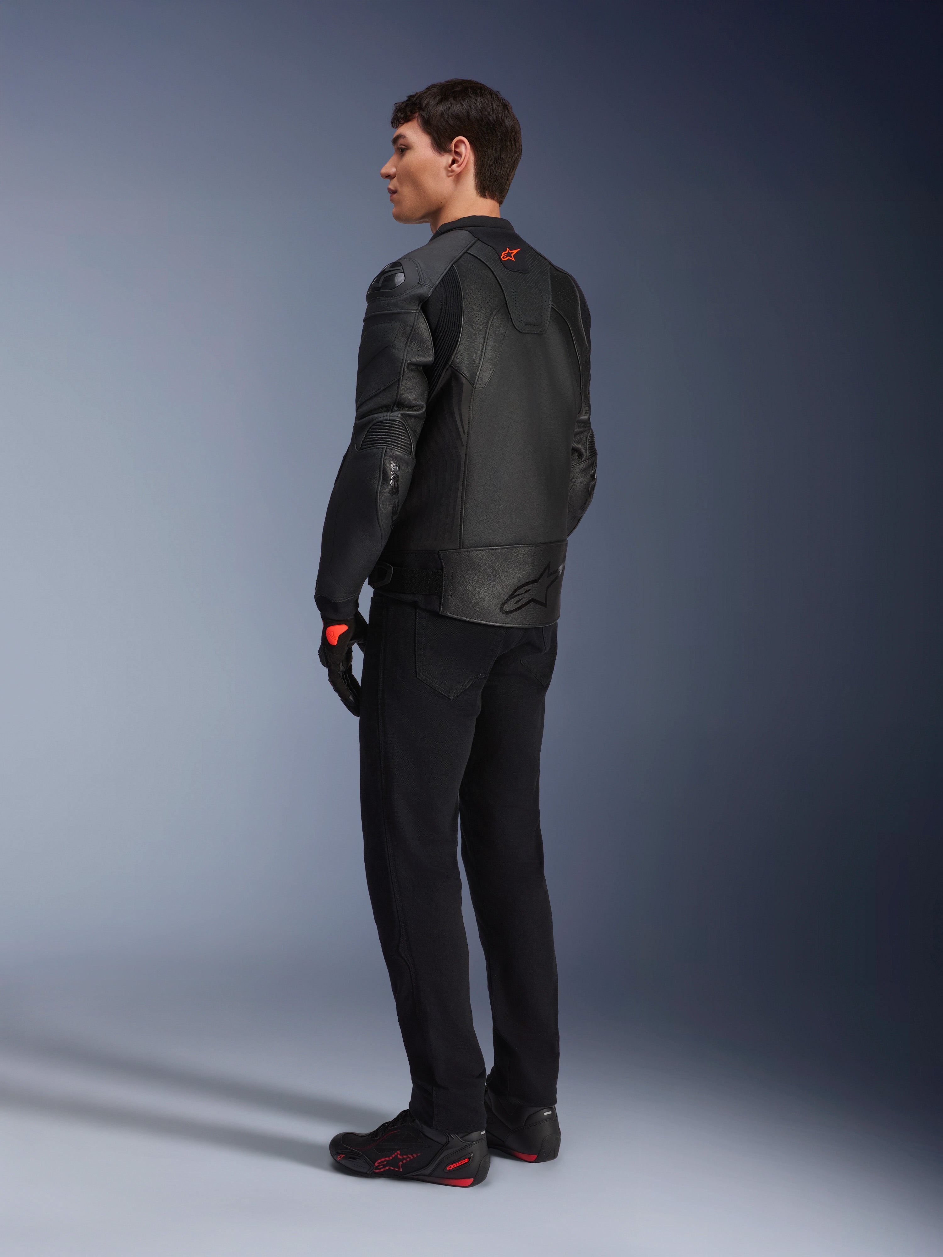 Chaqueta de cuero Alpinestars GP Plus R V4 Airflow, chaqueta deportiva de moto para hombre, totalmente negra, vista trasera de tres cuartos en un modelo, con paneles de cuero perforados, logotipo rojo en la espalda y protección de hombros integrada, combinada con pantalones negros y calzado de conducción