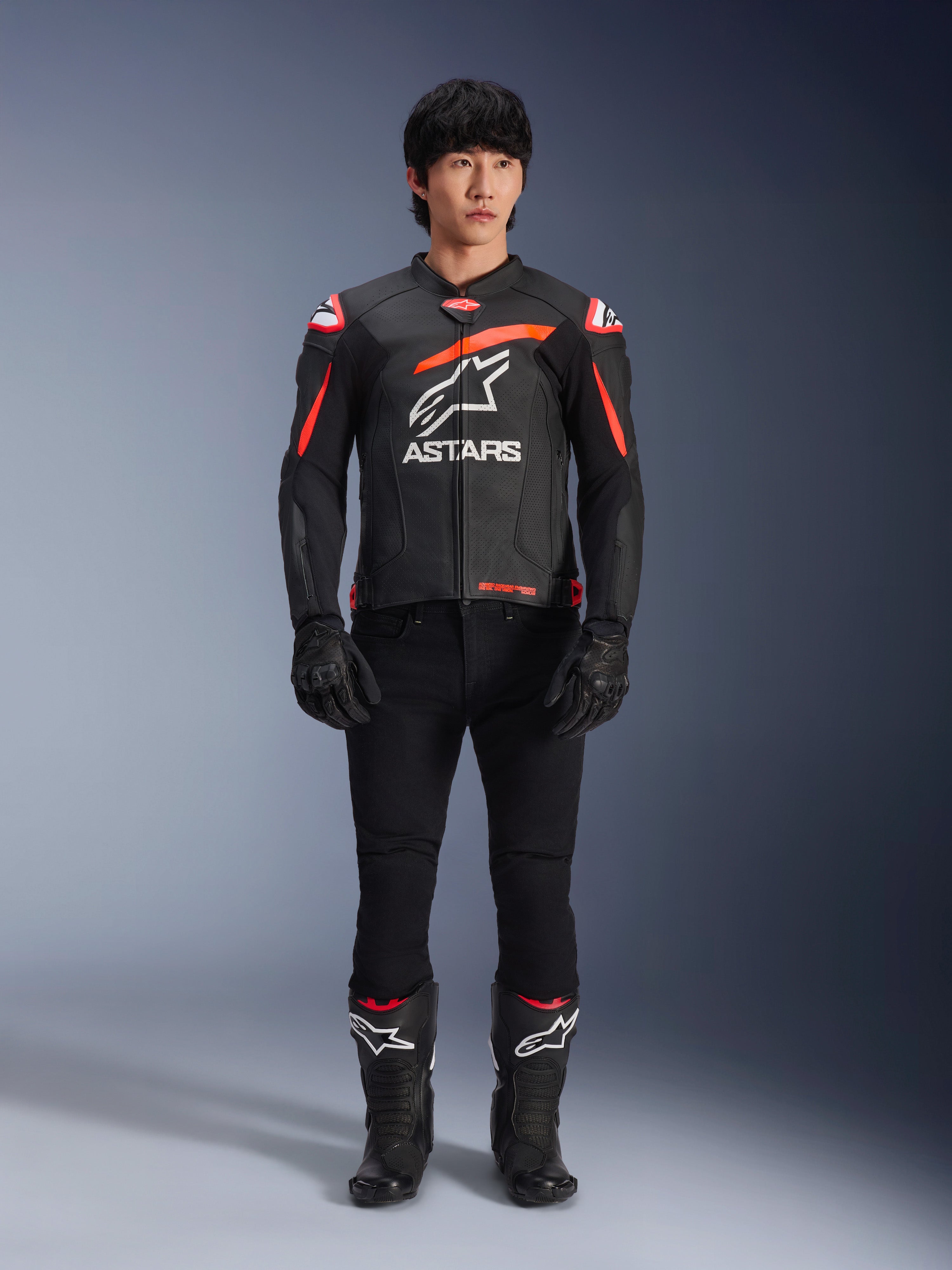 Modelo masculino con la chaqueta de cuero Alpinestars GP Plus R V4 Airflow, negra con detalles en rojo flúor y blanco, gran logotipo blanco de Astars en el pecho, acompañada de pantalones técnicos, guantes y botas negros, equipamiento de motociclismo de alto rendimiento en un estudio