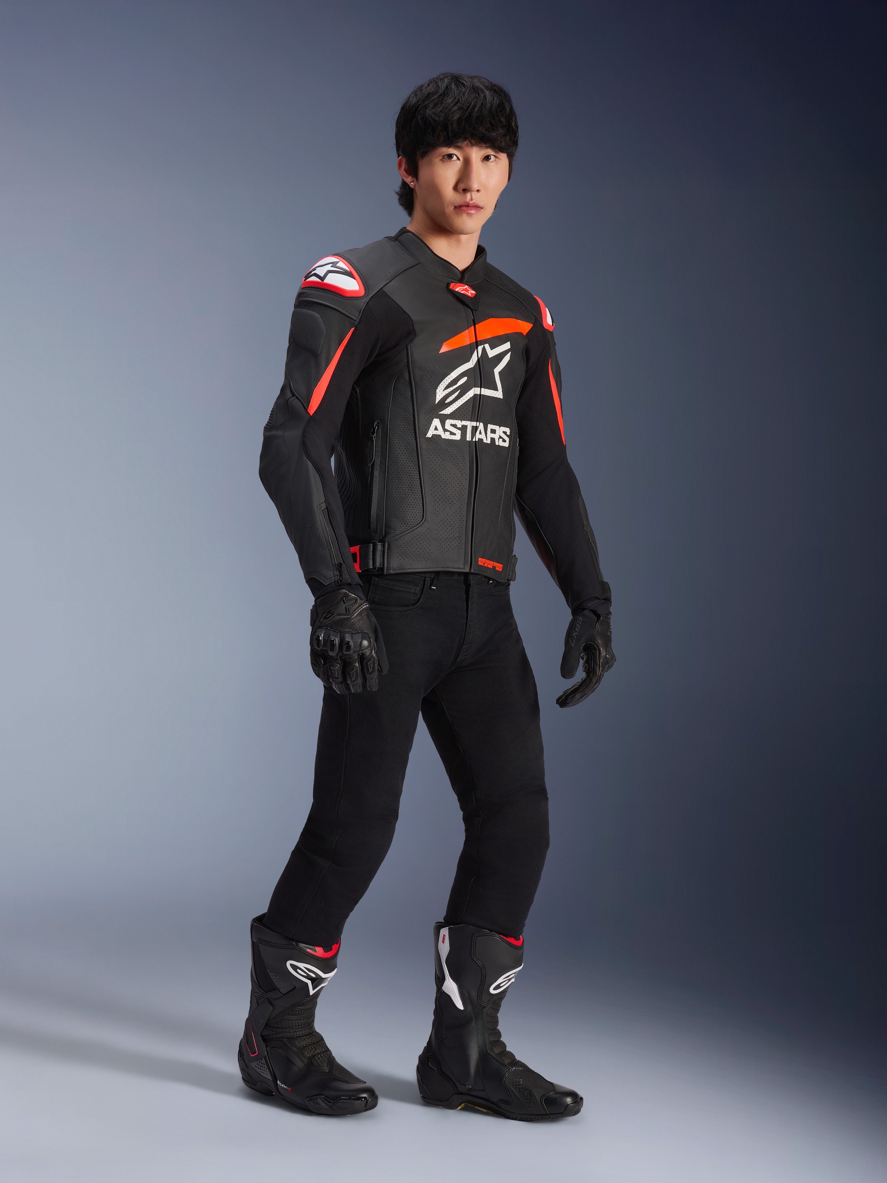 Piloto masculino con chaqueta de cuero Alpinestars GP Plus R V4 Airflow, negra con detalles en rojo flúor y blanco, con cuero perforado y logotipo ASTARS en el pecho, usada con pantalones de moto negros y botas de moto negras, equipamiento deportivo de alto rendimiento