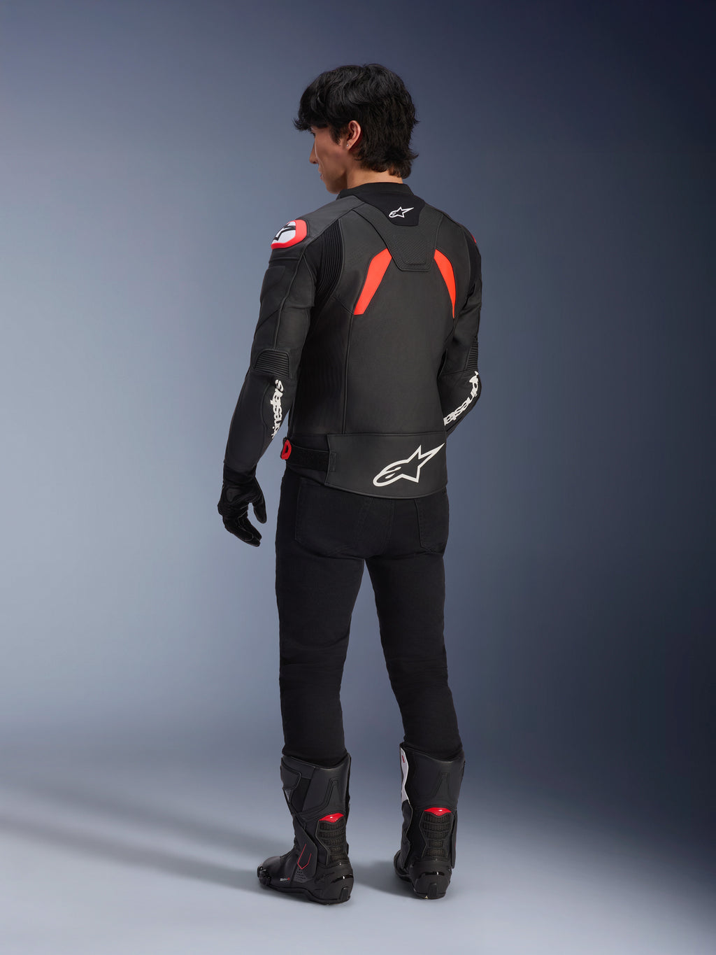 Vista trasera de un modelo con la chaqueta de cuero Alpinestars GP Plus R V4 Airflow, negro con detalles en rojo flúor y blanco, combinada con pantalones negros y botas de moto negras, con logotipos blancos prominentes en la espalda y las mangas, construcción de cuero de estilo deportivo