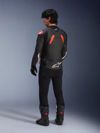 Vista trasera de un modelo con la chaqueta de cuero Alpinestars GP Plus R V4 Airflow, negro con detalles en rojo flúor y blanco, combinada con pantalones negros y botas de moto negras, con logotipos blancos prominentes en la espalda y las mangas, construcción de cuero de estilo deportivo