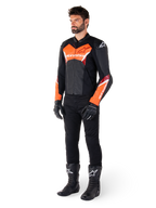 Piloto masculino con la chaqueta de cuero Alpinestars Faster V3 en negro y rojo flúor, combinada con pantalones de conducción, guantes y botas negros. La chaqueta presenta una silueta derivada de la competición con llamativos detalles en rojo flúor en el pecho y las mangas, logotipos de Alpinestars en blanco y una confección en cuero premium.