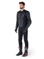 Hombre vistiendo la chaqueta de cuero Alpinestars Faster V3 Airflow en color negro, combinada con pantalones negros, guantes de moto negros y rojos, y botas de competición negras con detalles rojos y blancos, equipamiento de conducción deportiva de cuerpo completo