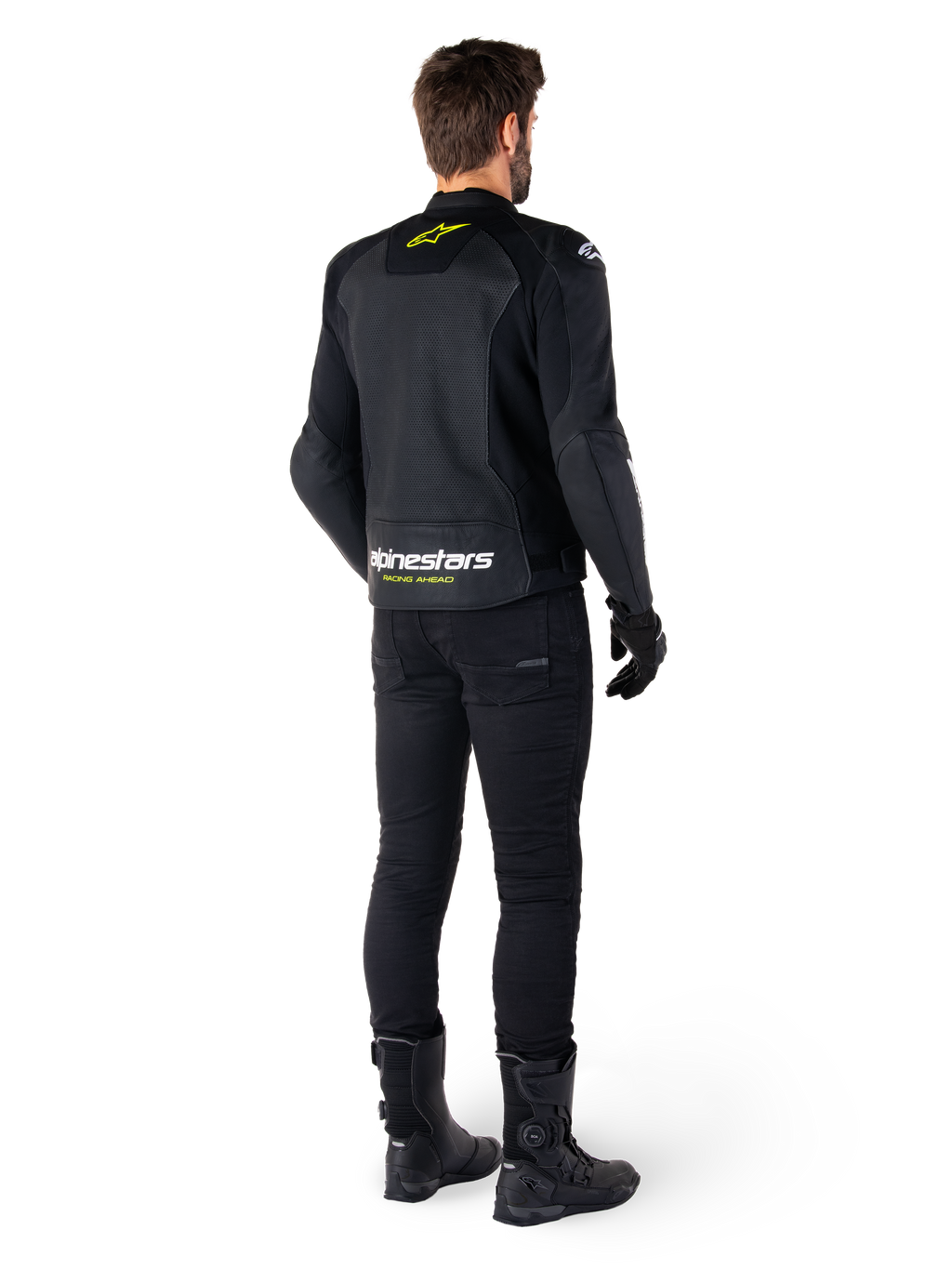 Vista trasera de un piloto con la chaqueta de cuero Alpinestars Faster V3 Airflow, negra con detalles en blanco y amarillo flúor, combinada con pantalones y botas negras, con cuero perforado para ventilación y la llamativa inscripción 'RACING AHEAD' en la parte inferior de la espalda.