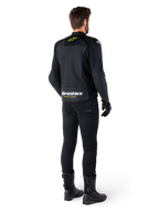 Vista trasera de un piloto con la chaqueta de cuero Alpinestars Faster V3 Airflow, negra con detalles en blanco y amarillo flúor, combinada con pantalones y botas negras, con cuero perforado para ventilación y la llamativa inscripción 'RACING AHEAD' en la parte inferior de la espalda.