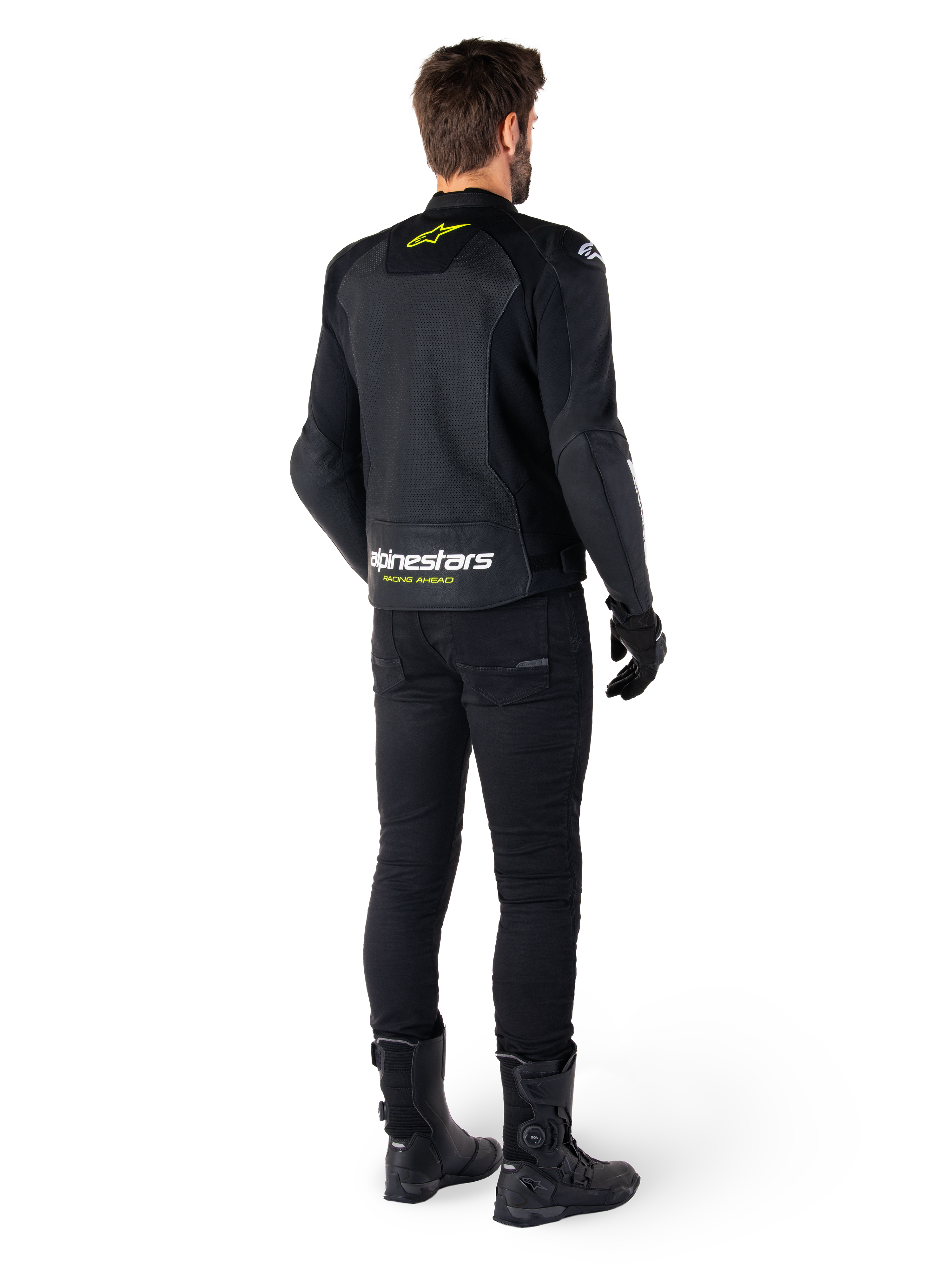 Vista trasera de un piloto con la chaqueta de cuero Alpinestars Faster V3 Airflow, negra con detalles en blanco y amarillo flúor, combinada con pantalones y botas negras, con cuero perforado para ventilación y la llamativa inscripción 'RACING AHEAD' en la parte inferior de la espalda.