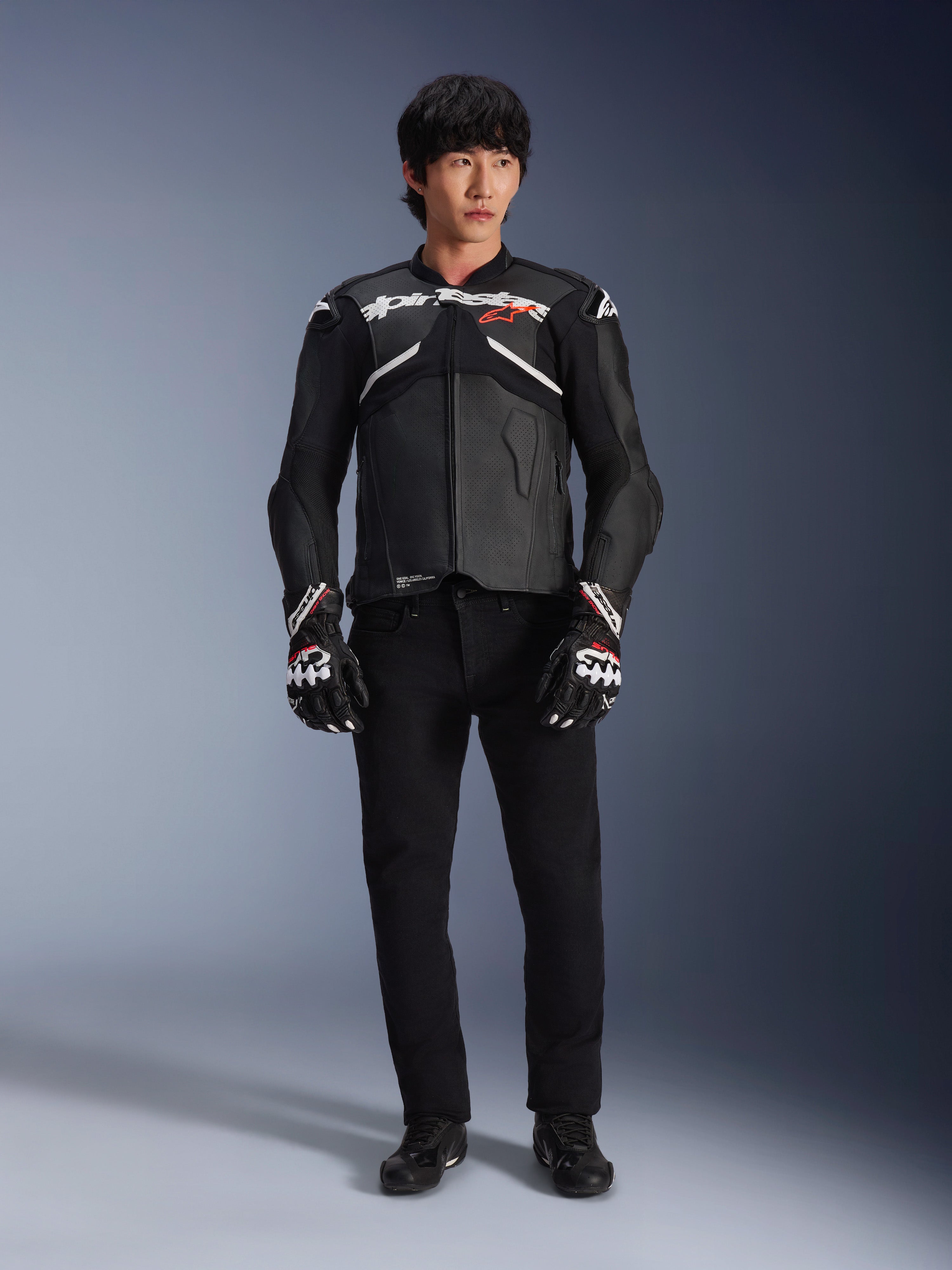 Modelo con la chaqueta de cuero Alpinestars Atem V5 en negro y blanco, con la marca Alpinestars en blanco y un logotipo de estrella roja en el pecho, combinada con pantalones negros y guantes de carreras en blanco y negro, equipamiento de motociclismo de alto rendimiento