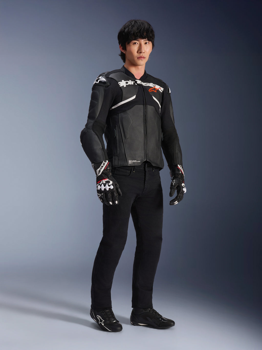 Chaqueta de cuero Alpinestars Atem V5, chaqueta de carreras para moto, negra y blanca, usada con pantalones negros y guantes a juego, con la marca Alpinestars en blanco sobre el pecho y protecciones en los hombros