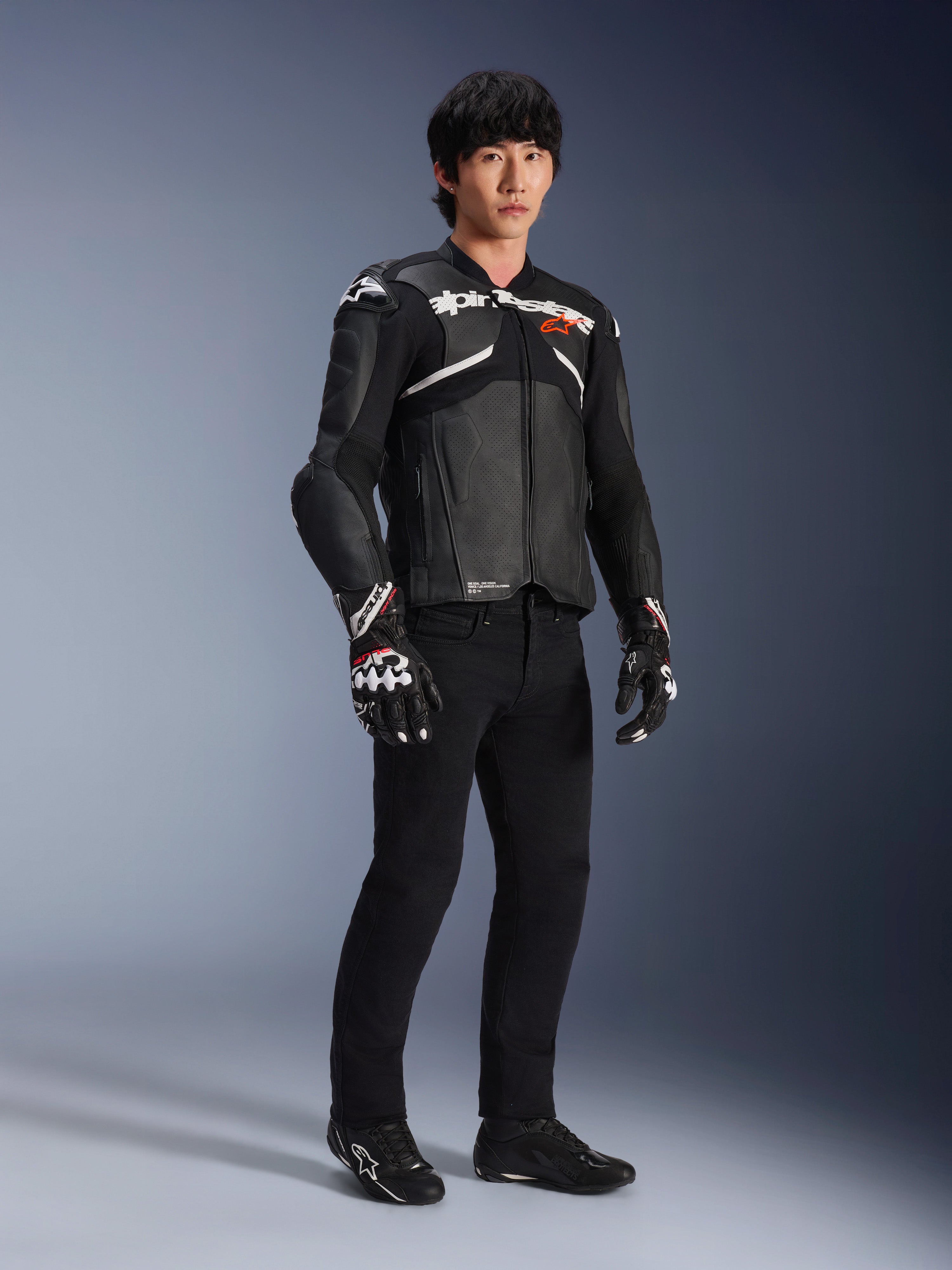 Chaqueta de cuero Alpinestars Atem V5, chaqueta de carreras para moto, negra y blanca, usada con pantalones negros y guantes a juego, con la marca Alpinestars en blanco sobre el pecho y protecciones en los hombros