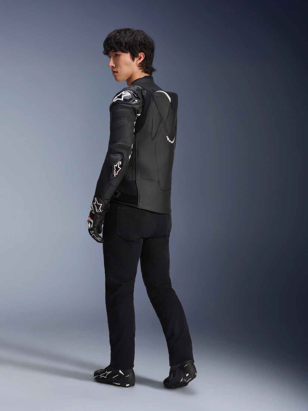 Modelo masculino con la chaqueta de cuero Alpinestars Atem V5 en negro y blanco, vista trasera de tres cuartos que muestra la joroba aerodinámica y los protectores DFS RR, combinada con pantalones negros, guantes de carreras y botas, equipamiento profesional de motociclismo