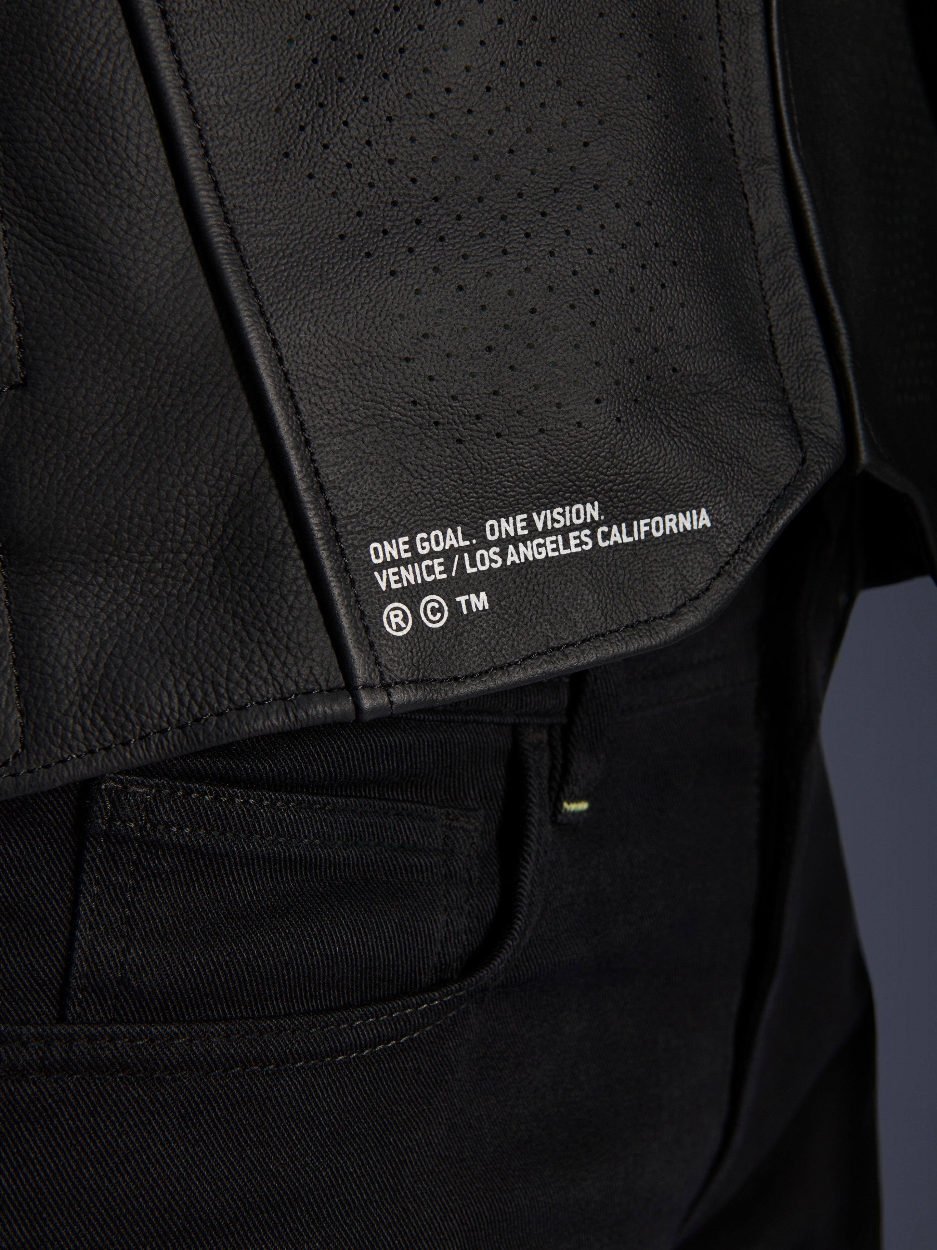 Primer plano de la chaqueta de cuero Alpinestars Atem V5 en color negro, con el panel inferior de la espalda que incluye la inscripción 'ONE GOAL. ONE VISION. VENICE / LOS ANGELES CALIFORNIA' y textura de cuero perforado.