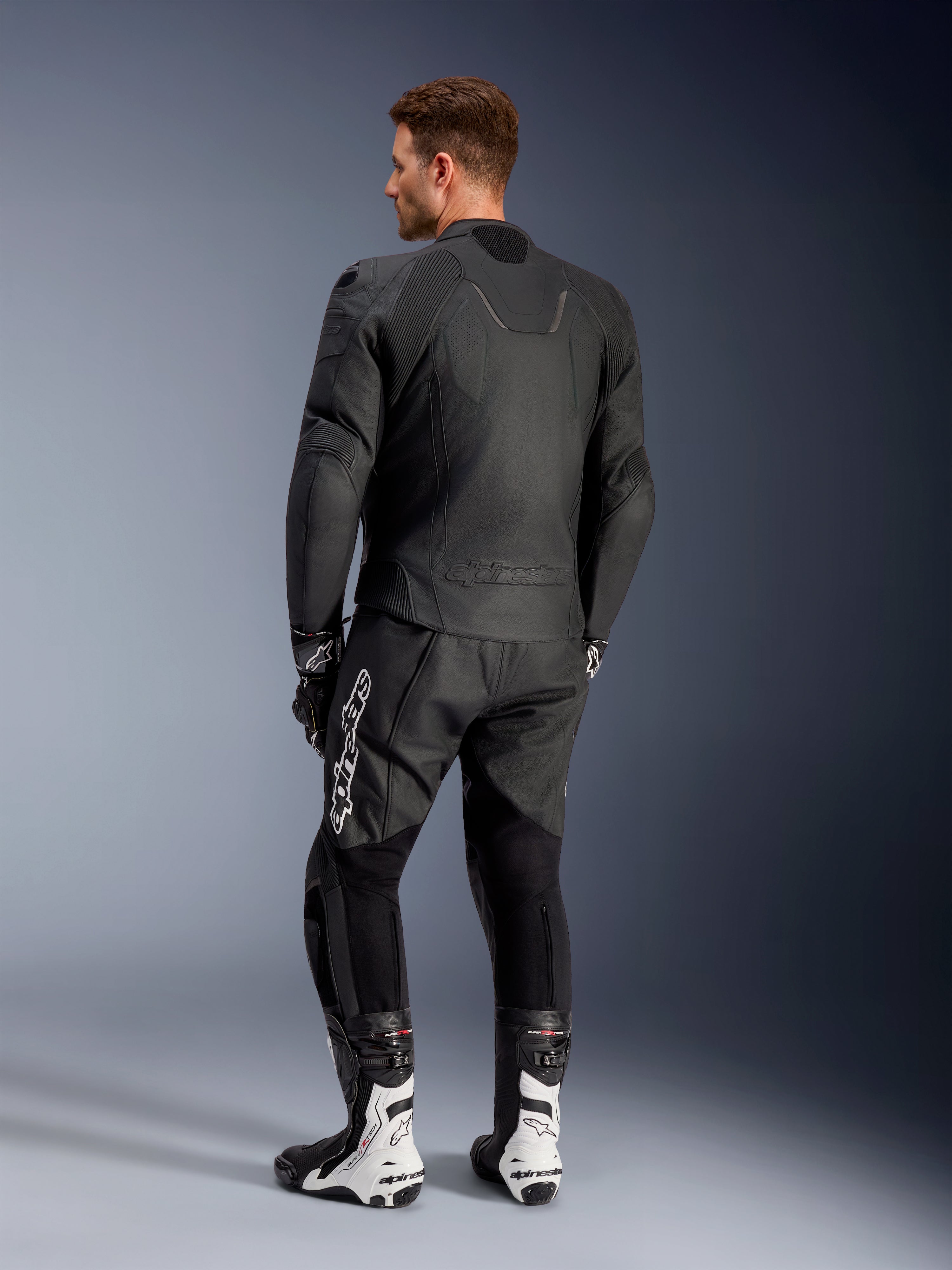 Alpinestars Caliber Leather Jacket, Black motorcycle road jacket, back view, featuring perforated leather and accordion stretch inserts on the waist and shoulders, embossed logo, worn with black leather pants and white racing boots