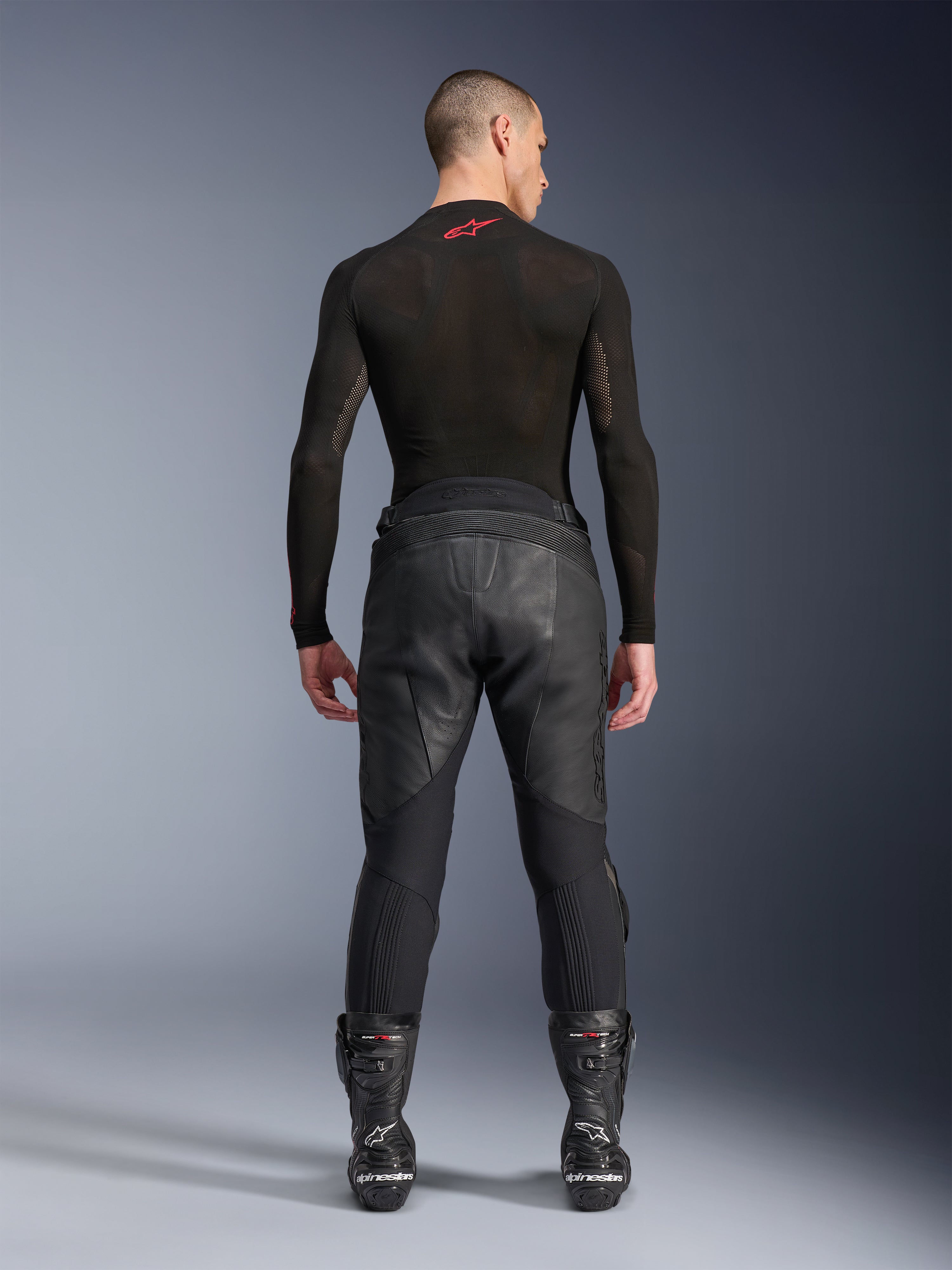 Alpinestars Missile V3 Leather Pants, negros, vista posterior en un modelo con camiseta de compresión de manga larga y botas de carreras, confección en cuero de primera calidad con superposición en la cintura alta y zonas elásticas, equipamiento profesional para carreras de motos