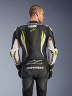 Mono de cuero de una pieza Alpinestars GP Tech V5, vista trasera en modelo, negro, blanco y amarillo flúor, con joroba aerodinámica, marca Alpinestars grande en los brazos y el asiento, paneles elásticos de acordeón y logo Tech-Air Ready