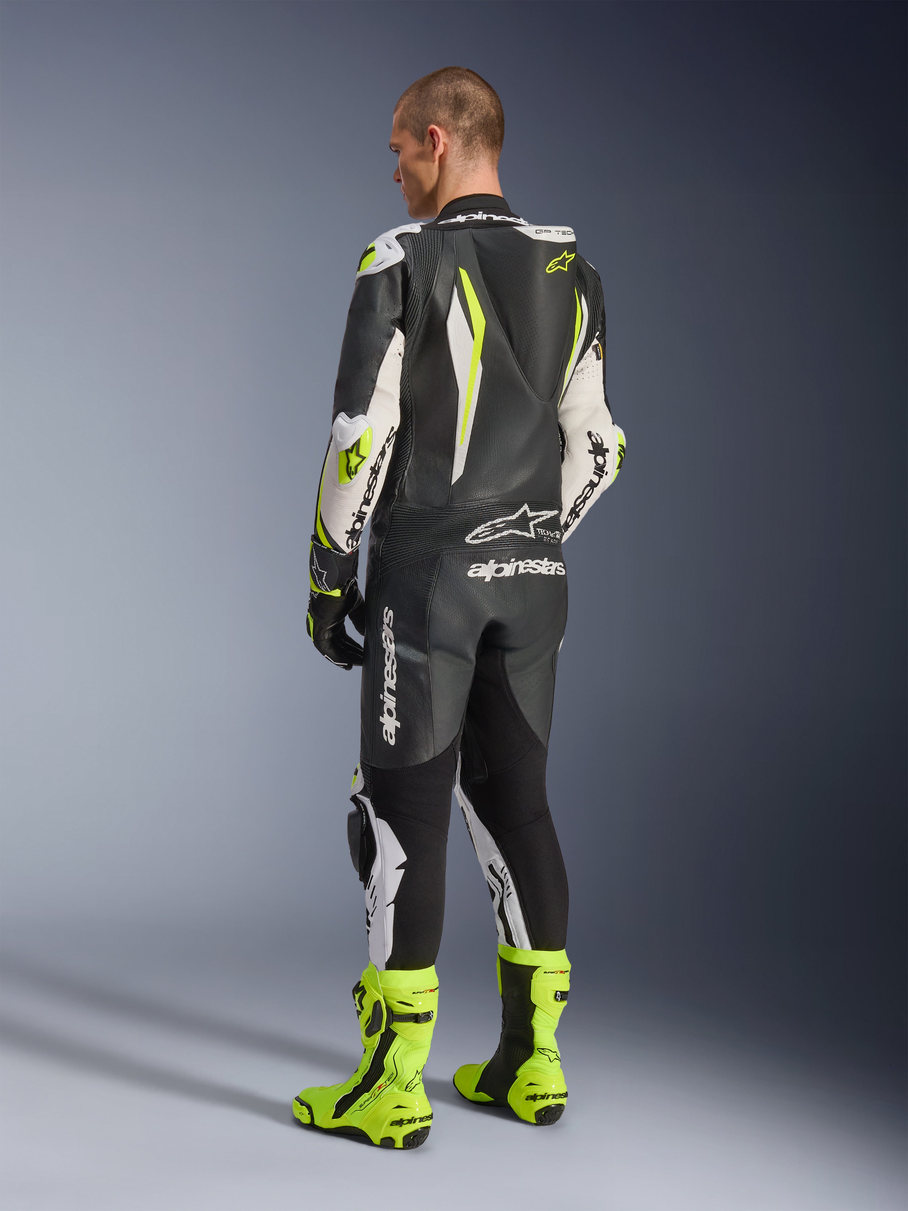 Mono de cuero de una pieza GP Tech V5, negro, blanco y amarillo flúor, vista trasera del piloto con el traje de carreras completo y botas amarillo flúor a juego, con joroba aerodinámica y logotipos de Alpinestars destacados, equipamiento profesional de motociclismo