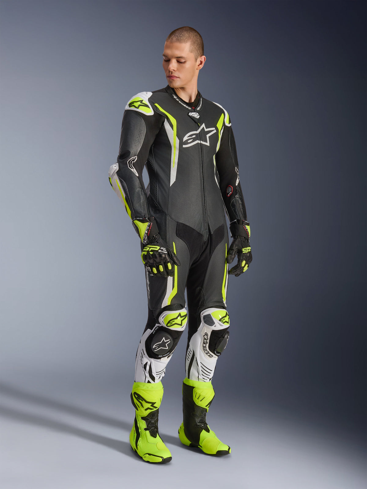 Alpinestars GP Tech V5 One Piece Leather Suit, negro, blanco y amarillo flúor, usado por un modelo con guantes y botas de carreras a juego, equipamiento profesional para carreras de motos con rendimiento aerodinámico y protecciones.