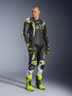 Alpinestars GP Tech V5 One Piece Leather Suit, negro, blanco y amarillo flúor, usado por un modelo con guantes y botas de carreras a juego, equipamiento profesional para carreras de motos con rendimiento aerodinámico y protecciones.