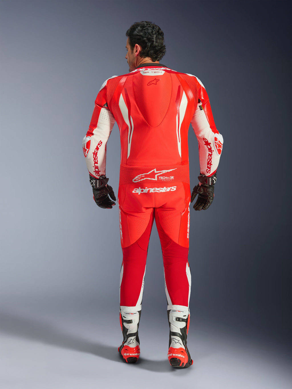 Mono de cuero de una pieza Alpinestars GP Tech V5, traje de carreras para motociclismo, rojo flúor, rojo brillante y blanco, vista trasera que muestra la joroba aerodinámica y el logotipo de Alpinestars en blanco, usado con guantes y botas de carreras en blanco y negro