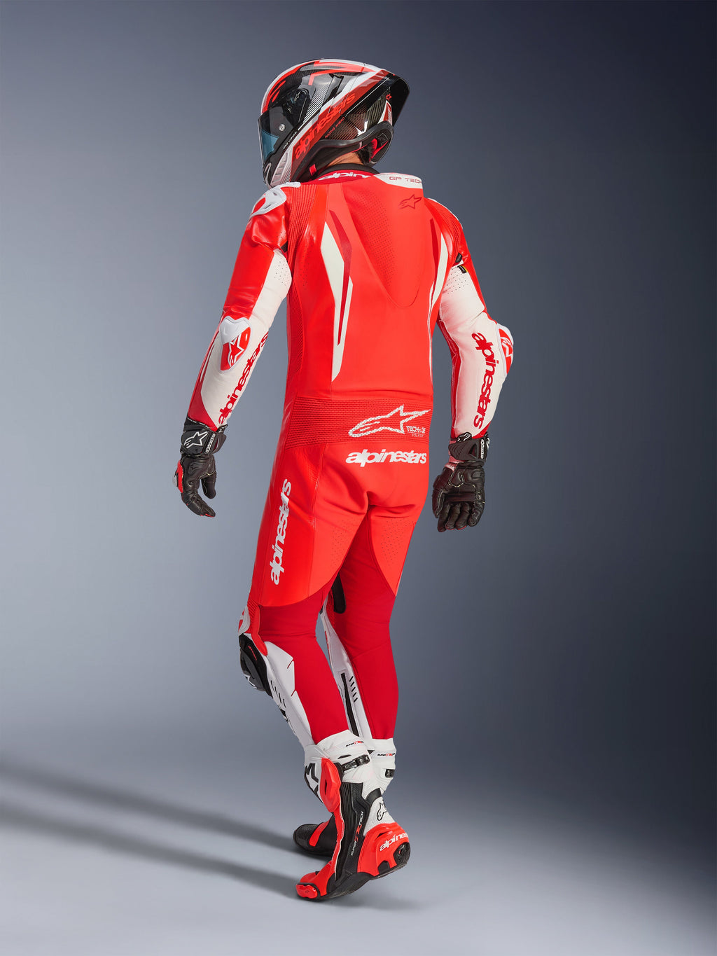 Alpinestars GP Tech V5 One Piece Leather Suit, mono de cuero de una pieza en rojo flúor, rojo brillante y blanco, vista trasera de un motociclista con equipamiento de carreras completo, con joroba aerodinámica y paneles blancos en mangas y piernas, combinado con casco a juego y botas de carreras rojas, diseño para rendimiento profesional en pista.