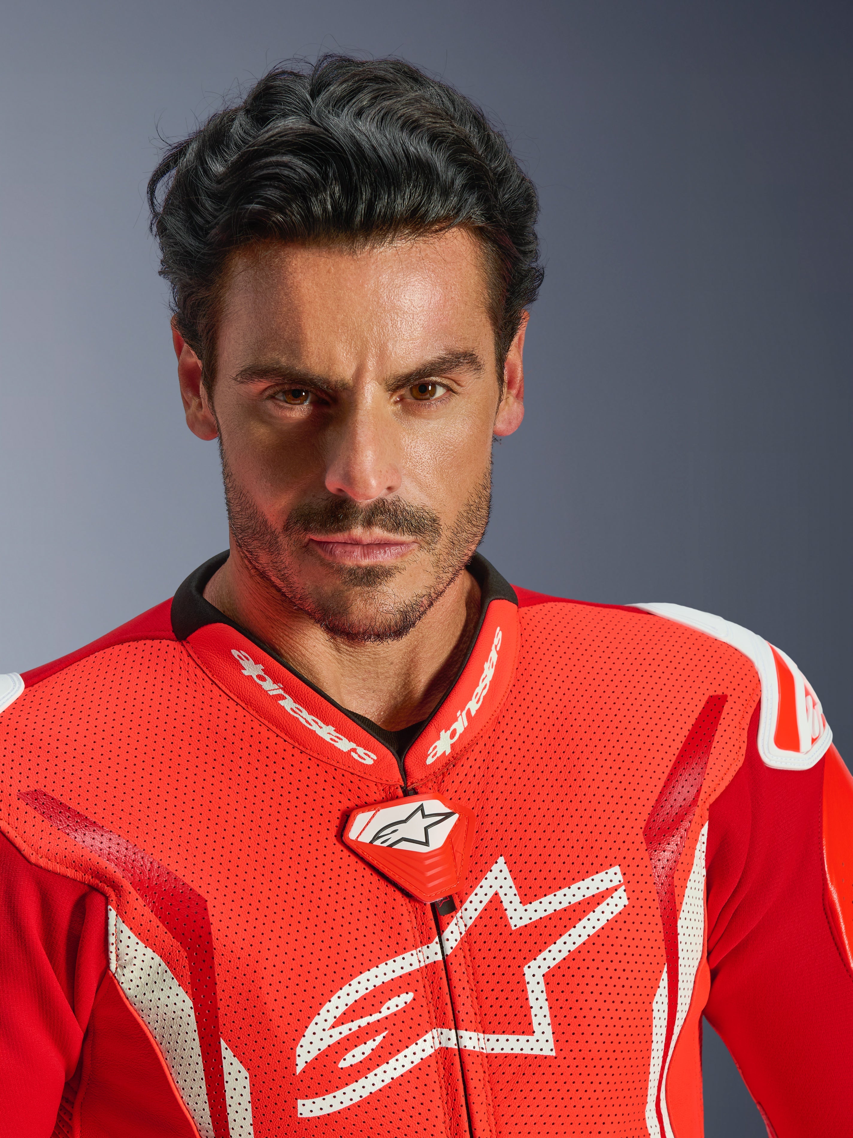 Mono de cuero de una pieza Alpinestars GP Tech V5, traje de competición para moto, Rojo Flúor, Rojo Brillante y Blanco, retrato en primer plano de un piloto, pecho de cuero perforado con logo de Alpinestars en blanco, cuello de competición de alto rendimiento y diseño aerodinámico.