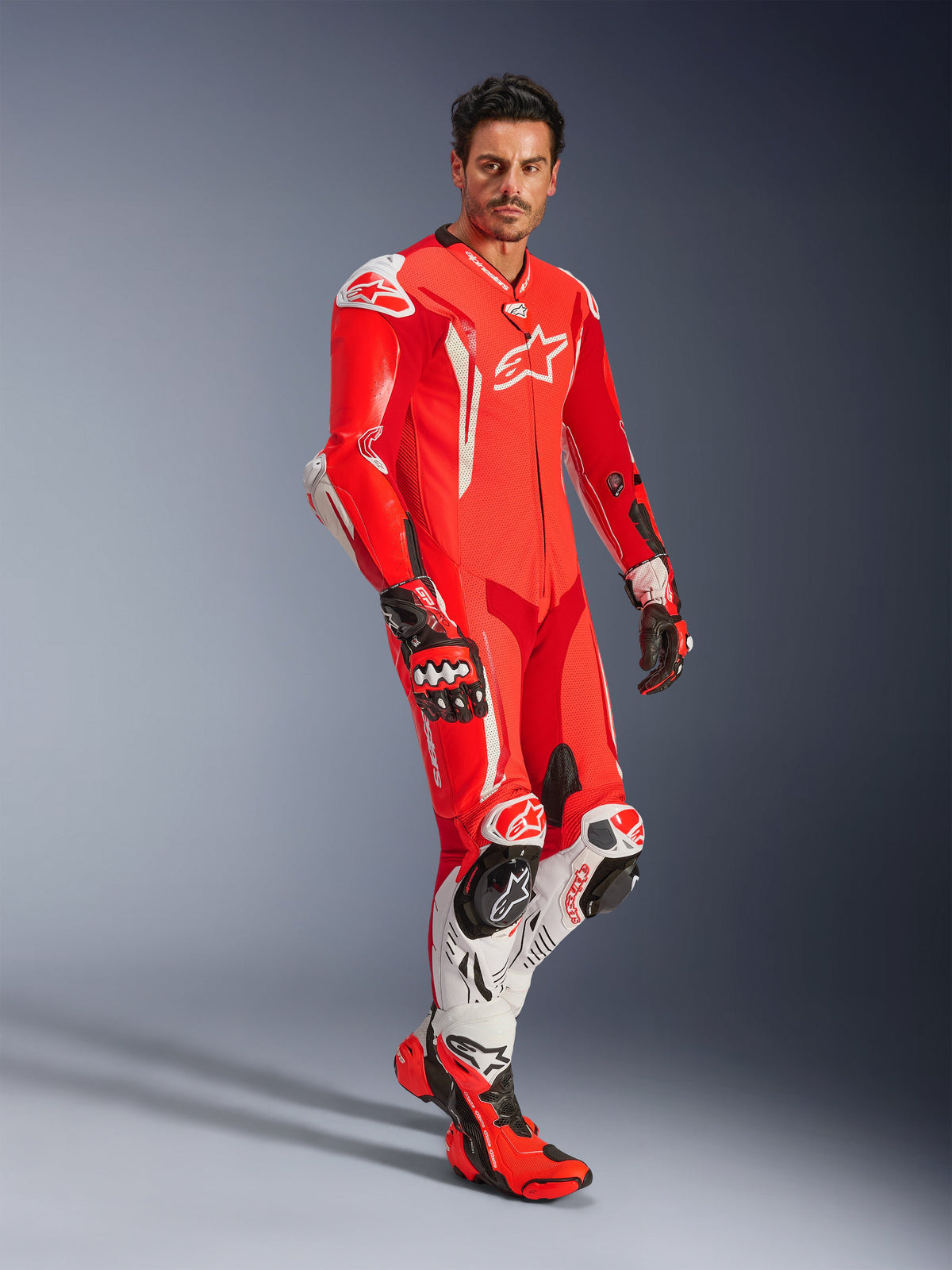 Hombre con el traje de cuero de una pieza Alpinestars GP Tech V5, en rojo flúor, rojo brillante y blanco, combinado con guantes y botas de competición, equipamiento profesional de carreras de motos con joroba aerodinámica y protección integrada.