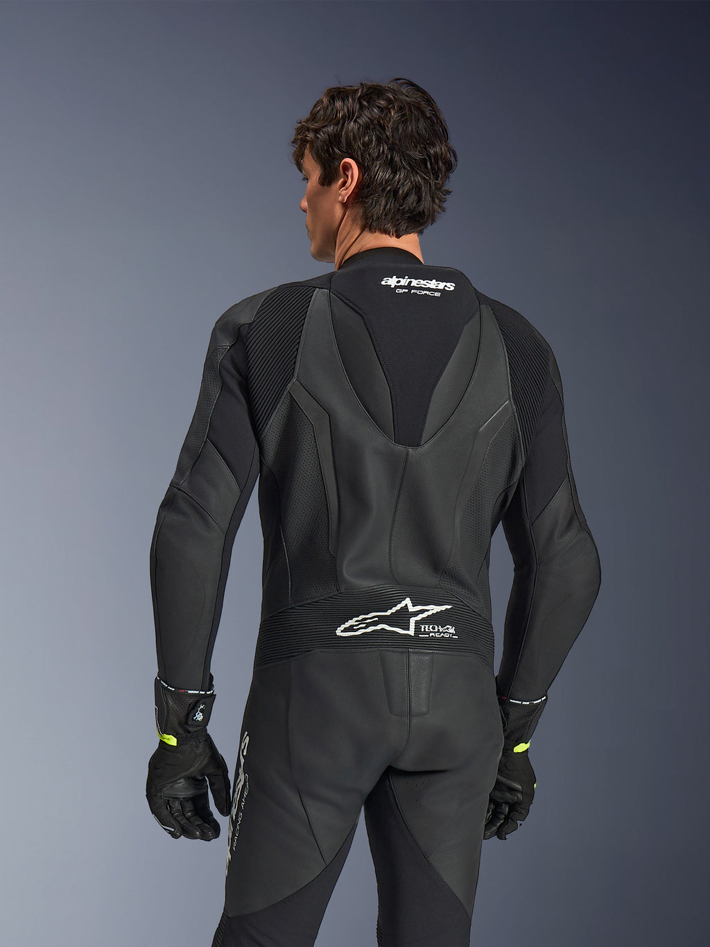 Alpinestars GP Force V2 Leather Suit, 1-Piece, Black, back view showing perforated leather and accordion stretch panels, white Alpinestars and Tech-Air Ready branding on the upper and lower back, worn with black gloves, high-performance motorcycle racing gear