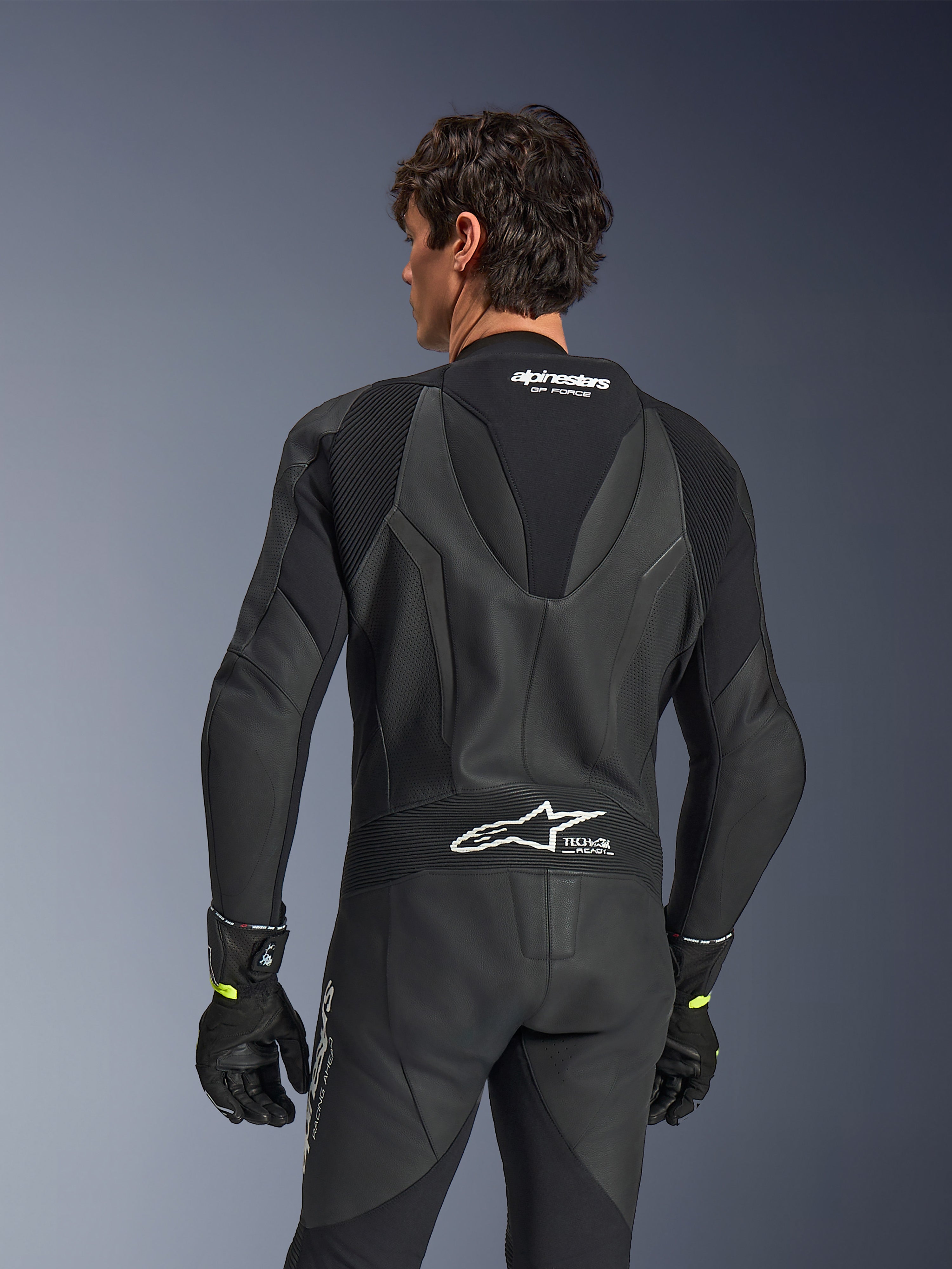 Alpinestars GP Force V2 Leather Suit, 1-Piece, Black, back view showing perforated leather and accordion stretch panels, white Alpinestars and Tech-Air Ready branding on the upper and lower back, worn with black gloves, high-performance motorcycle racing gear
