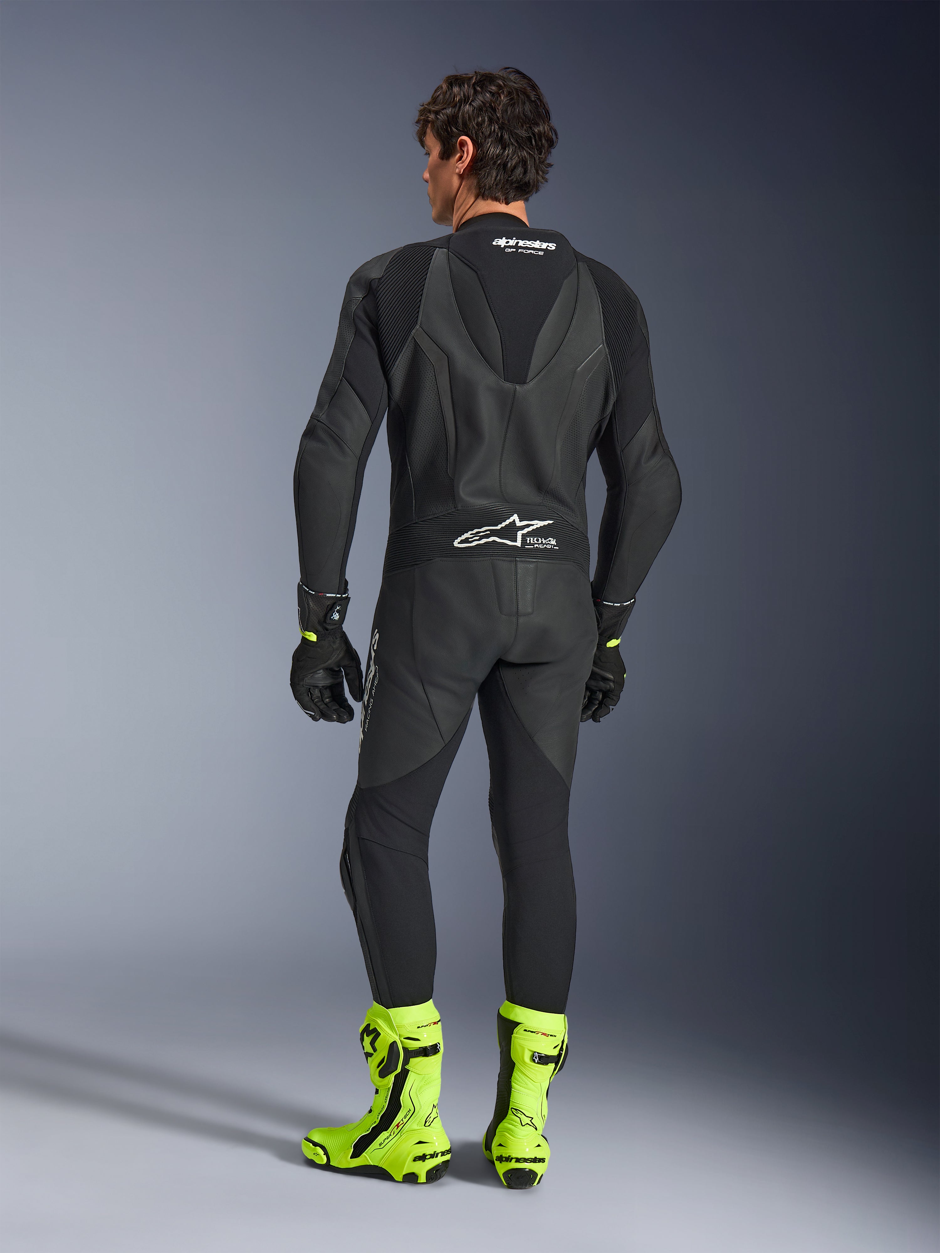 Alpinestars GP Force V2 Leather Suit - 1Piece, Black, back view worn by a model, featuring an aerodynamic speed hump and Tech-Air Ready branding, styled with black gloves and bright fluorescent yellow racing boots, professional track equipment