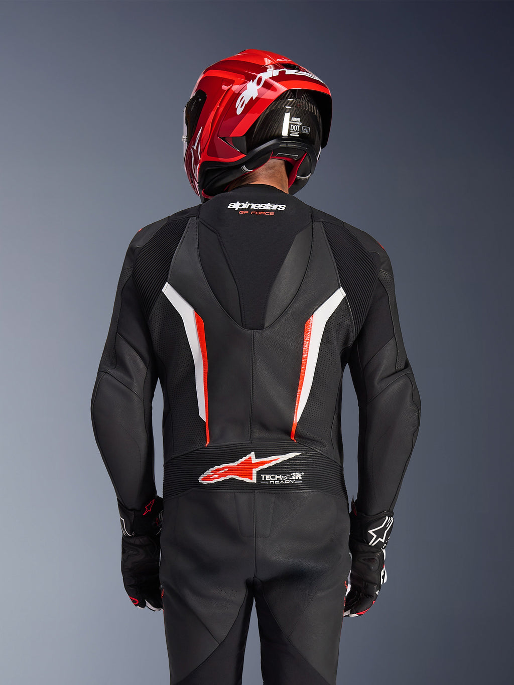 Traje de cuero de una pieza Alpinestars GP Force V2, negro, blanco y rojo fluo, vista posterior de un piloto con casco de carreras rojo y guantes, con paneles de cuero perforado, joroba aerodinámica con marca Alpinestars y logotipo Tech-Air Ready en la parte baja de la espalda, equipamiento profesional de motociclismo.