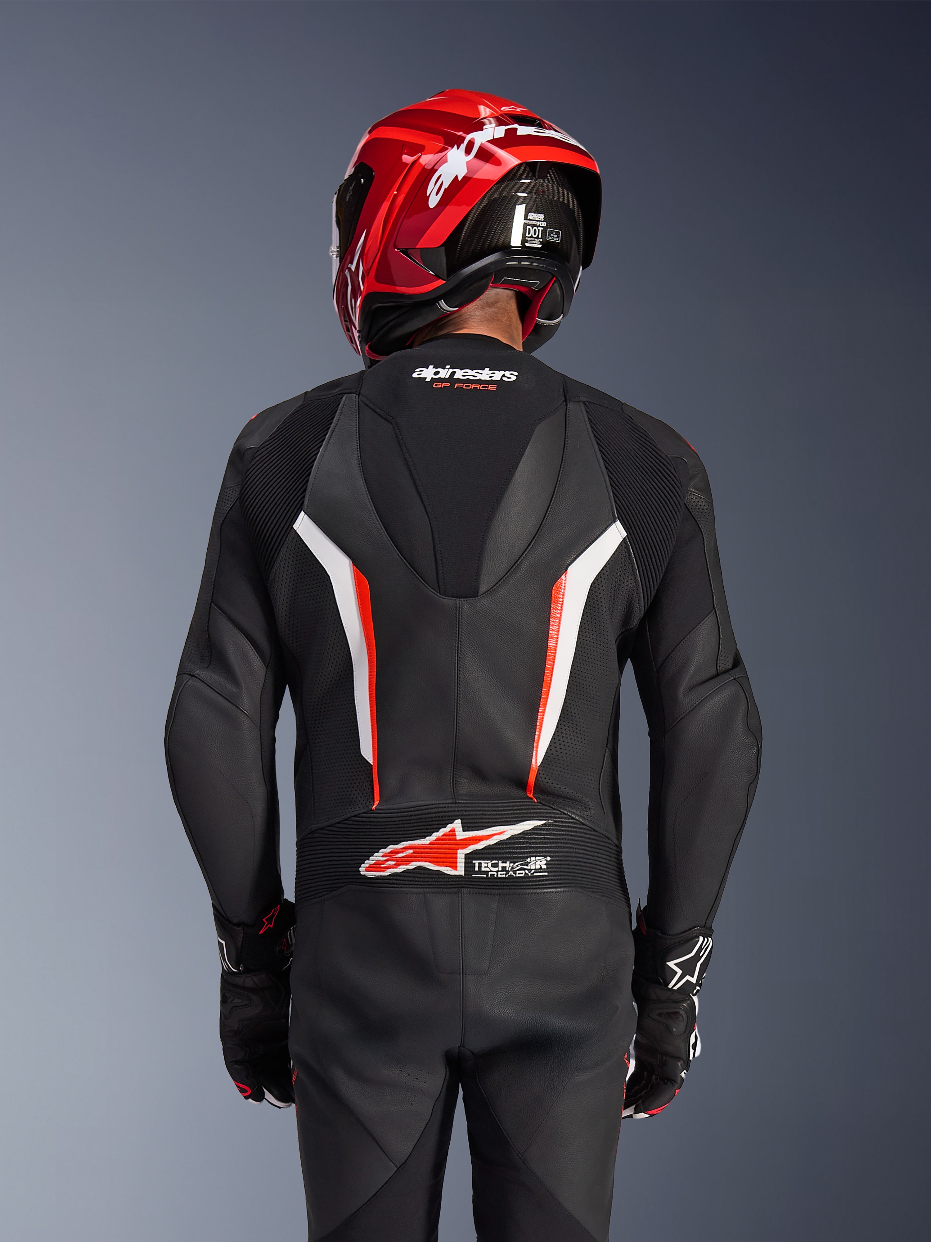 Traje de cuero de una pieza Alpinestars GP Force V2, negro, blanco y rojo fluo, vista posterior de un piloto con casco de carreras rojo y guantes, con paneles de cuero perforado, joroba aerodinámica con marca Alpinestars y logotipo Tech-Air Ready en la parte baja de la espalda, equipamiento profesional de motociclismo.