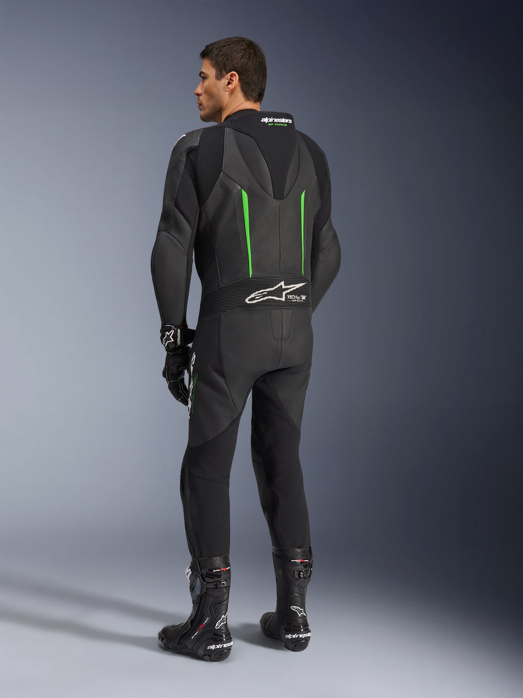 Traje de cuero Alpinestars GP Force V2, mono de motociclismo de una pieza, negro con detalles en verde, vista trasera que muestra la joroba aerodinámica y el logotipo blanco en el panel elástico de acordeón, combinado con botas y guantes de carreras negros.