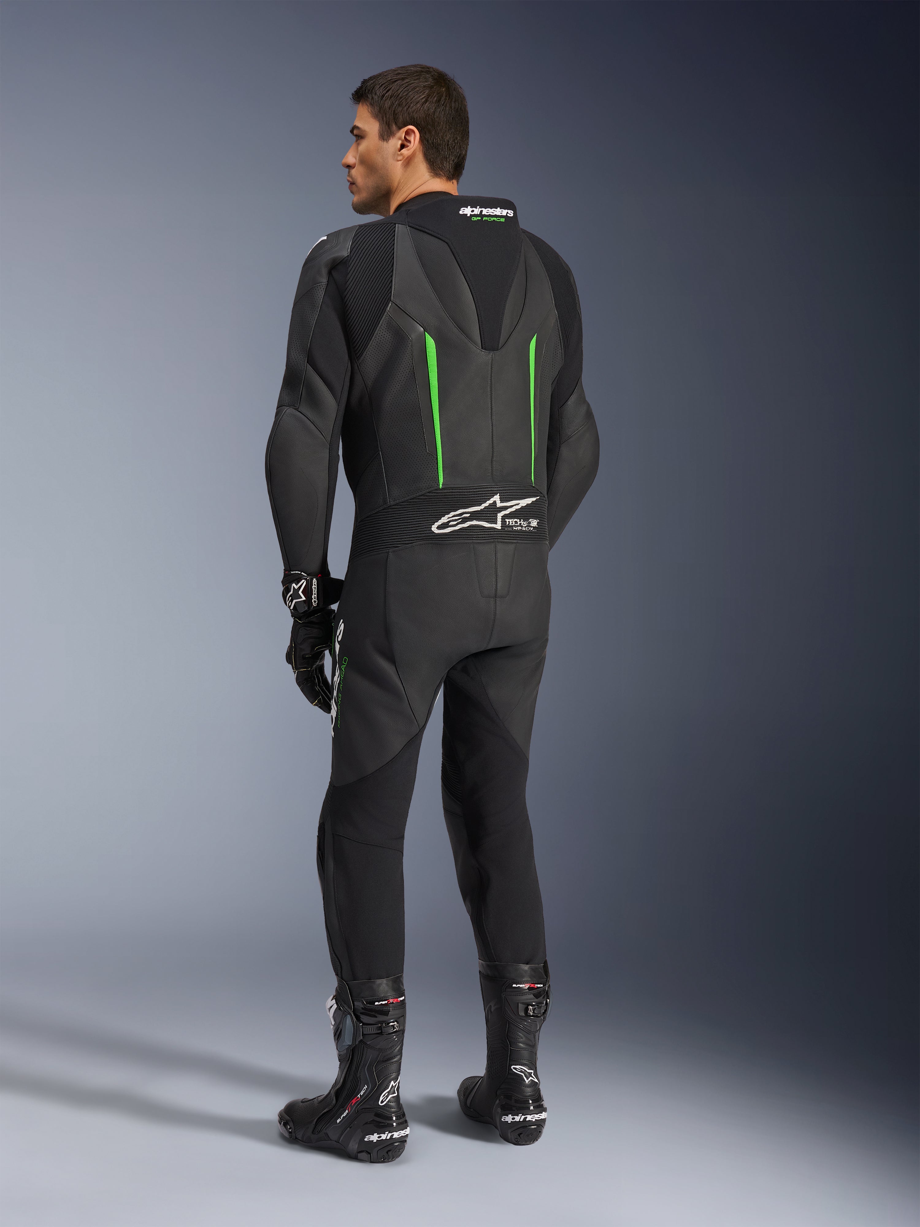 Traje de cuero Alpinestars GP Force V2, mono de motociclismo de una pieza, negro con detalles en verde, vista trasera que muestra la joroba aerodinámica y el logotipo blanco en el panel elástico de acordeón, combinado con botas y guantes de carreras negros.