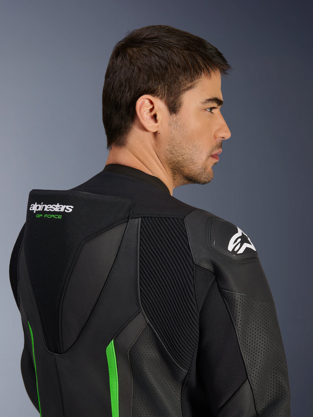 Primer plano de la parte superior de la espalda y el hombro de un modelo masculino que viste el traje de cuero Alpinestars GP Force V2 en negro y verde, con la joroba aerodinámica con la marca 'GP FORCE' en verde, un logotipo de Astars blanco en el hombro y paneles elásticos de acordeón.