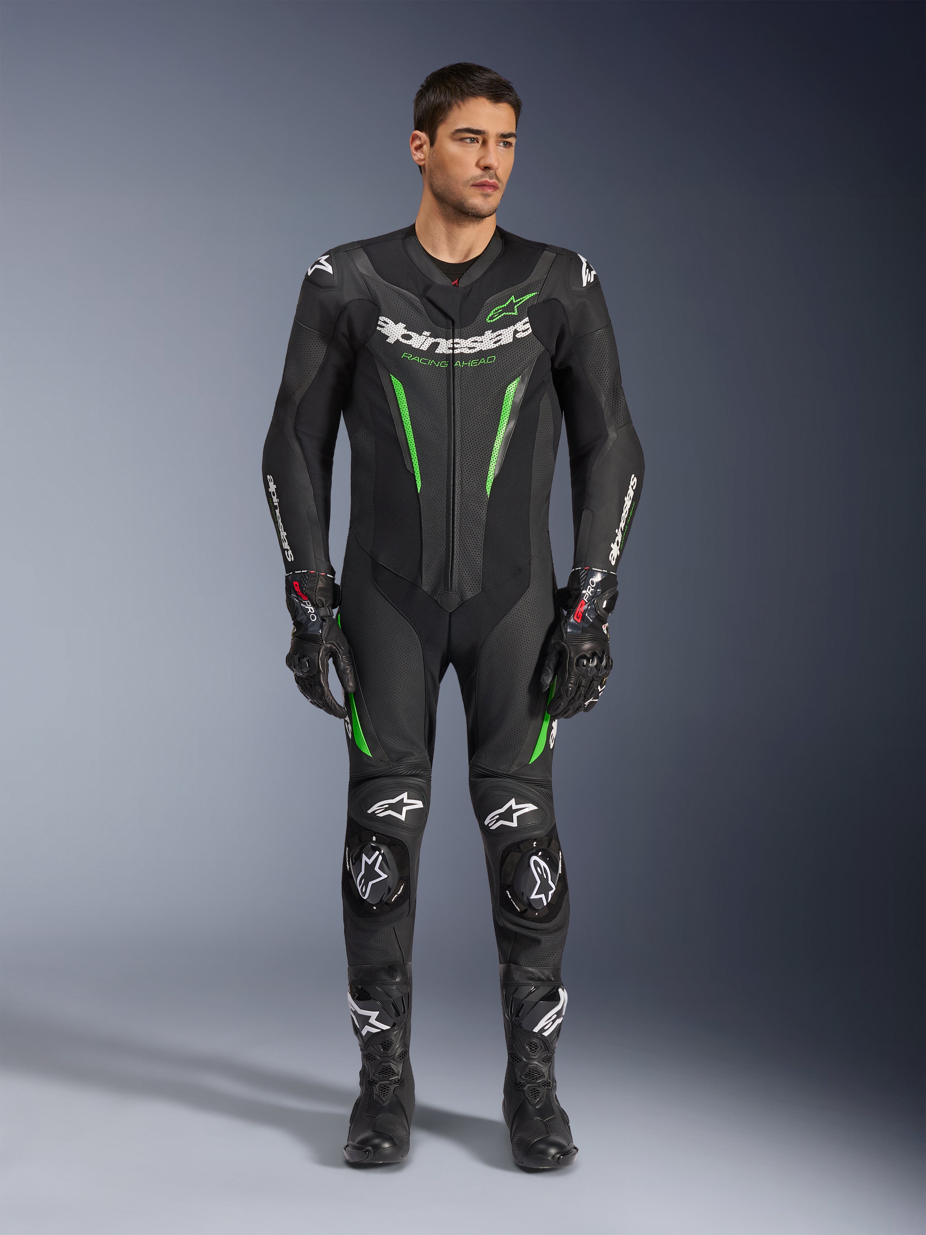 Alpinestars GP Force V2 Leather Suit - 1Piece, mono de competición de una pieza para motocicleta, negro y verde, vista frontal de un modelo vistiendo el traje con marca blanca en el pecho y detalles en verde neón, con cuero perforado y deslizadores externos en los hombros, combinado con guantes y botas de carreras negros