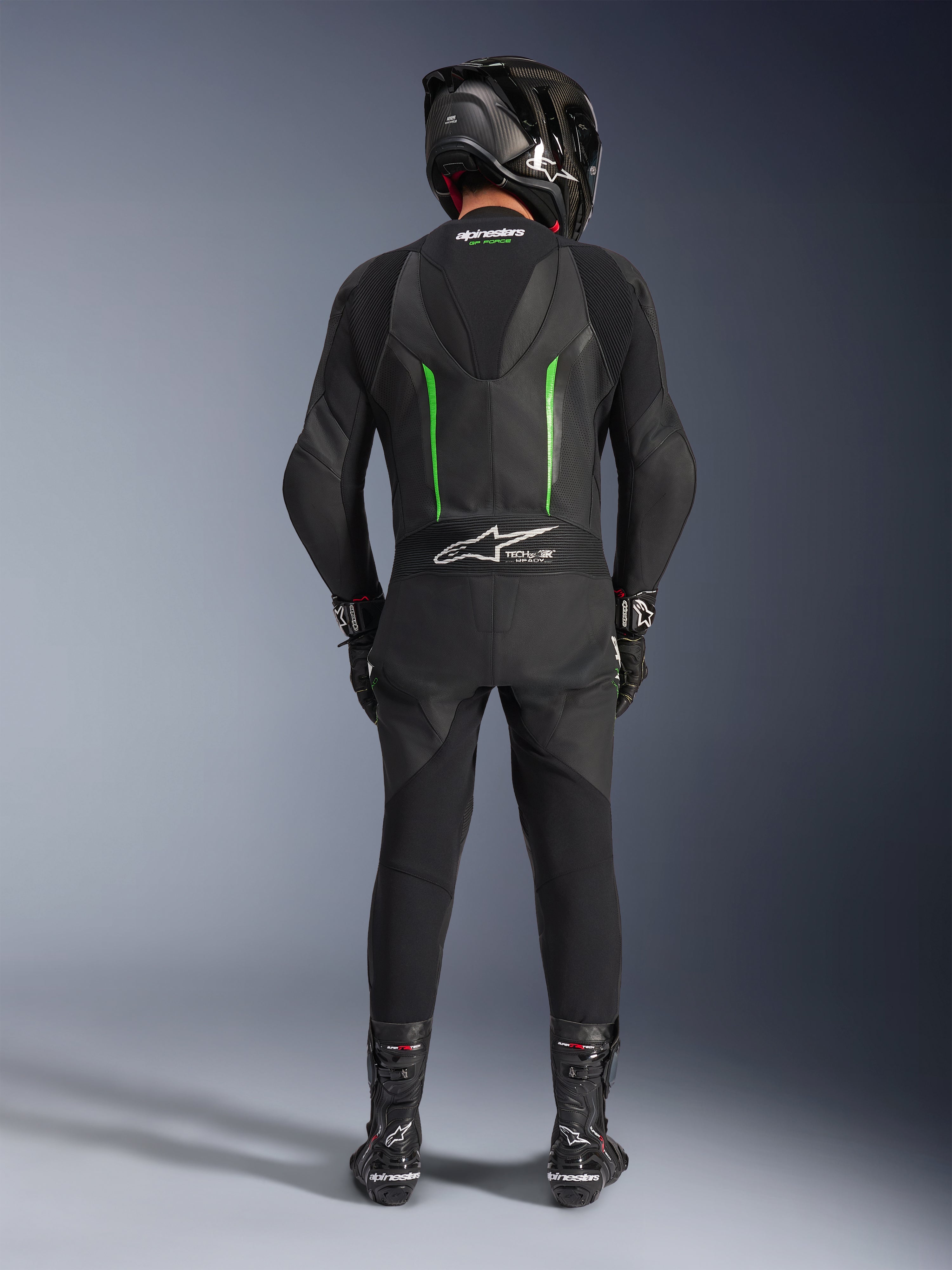Piloto visto desde atrás con el traje de cuero de una pieza Alpinestars GP Force V2 en negro y verde, combinado con un casco de fibra de carbono negro, guantes de carreras negros y botas negras, con una joroba aerodinámica y el logotipo Tech-Air Ready.