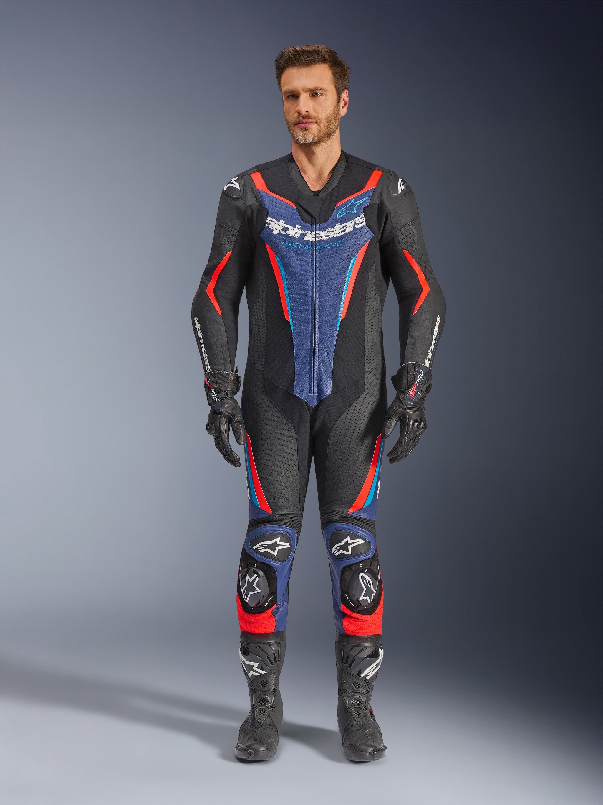 Modelo masculino con el traje de cuero Alpinestars GP Force V2 de 1 pieza en negro, azul y rojo flúor, vista frontal de cuerpo completo, incluye guantes y botas de carreras negros, con paneles de cuero perforado y logotipos distintivos de Alpinestars para rendimiento profesional en pista