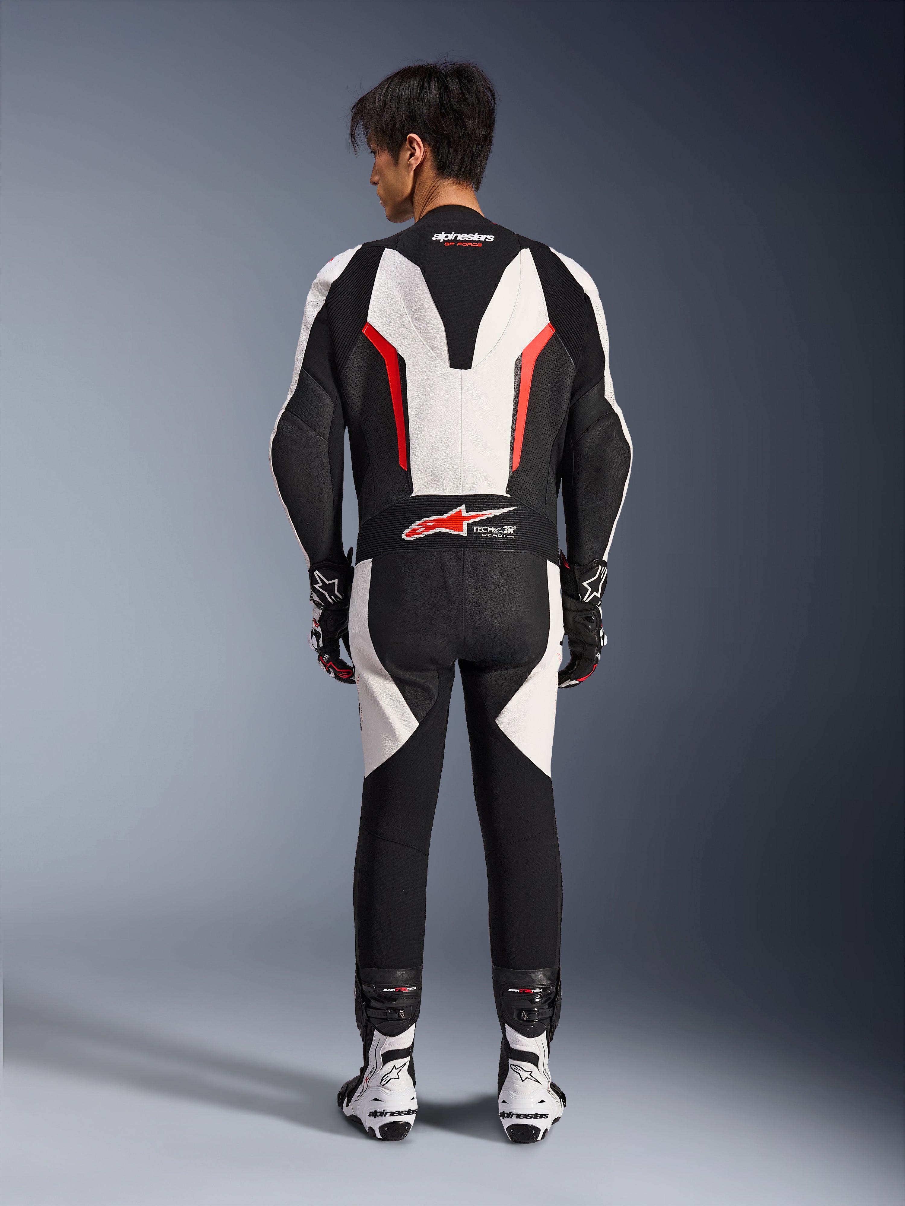 Traje de cuero Alpinestars GP Force V2 de 1 pieza, traje de carreras para moto, blanco, negro y rojo flúor, vista posterior del modelo usando el traje con botas y guantes de carreras, con joroba aerodinámica, paneles elásticos de acordeón y logotipo Tech-Air Ready