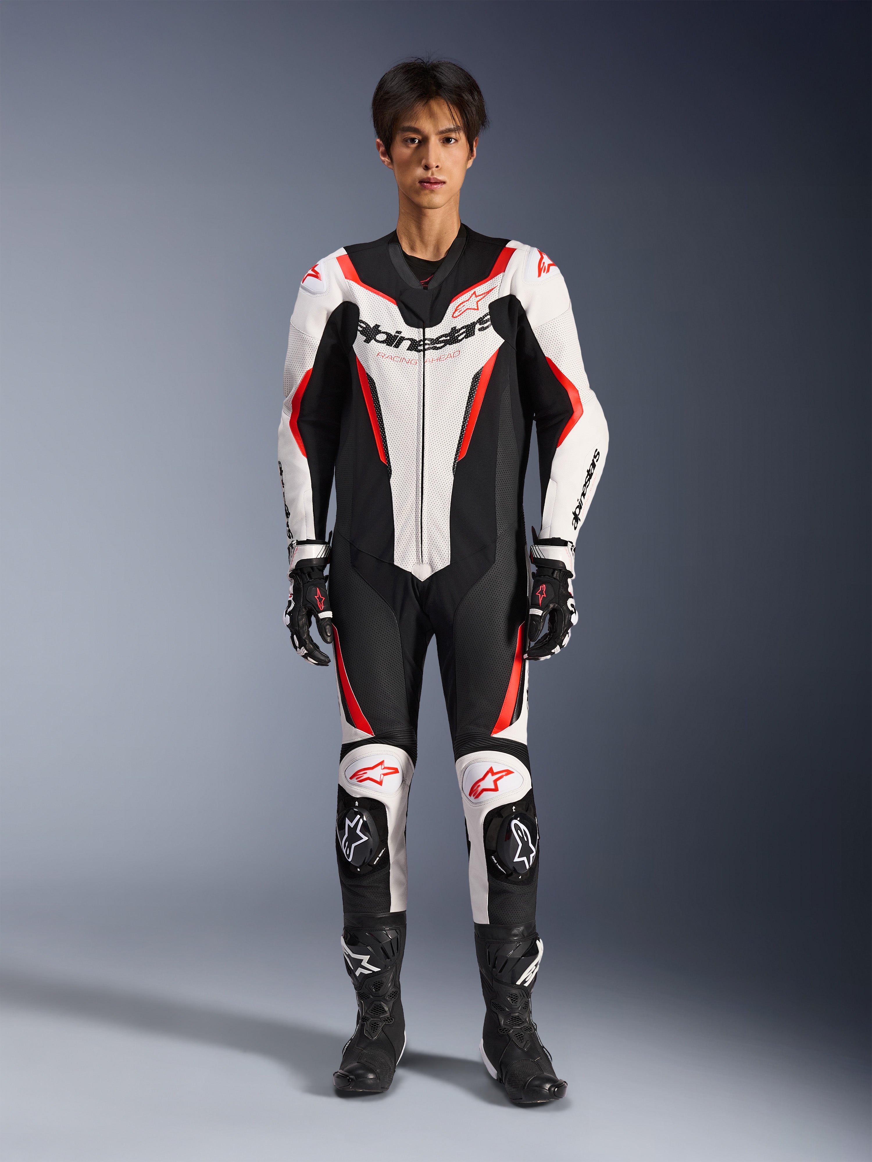 Traje de cuero Alpinestars GP Force V2 - 1 pieza, blanco negro rojo flúor, mono de cuero de competición de una pieza, vista frontal en modelo, con paneles perforados y detalles en rojo, logotipo de Alpinestars destacado en el pecho, combinado con botas y guantes de carreras profesionales