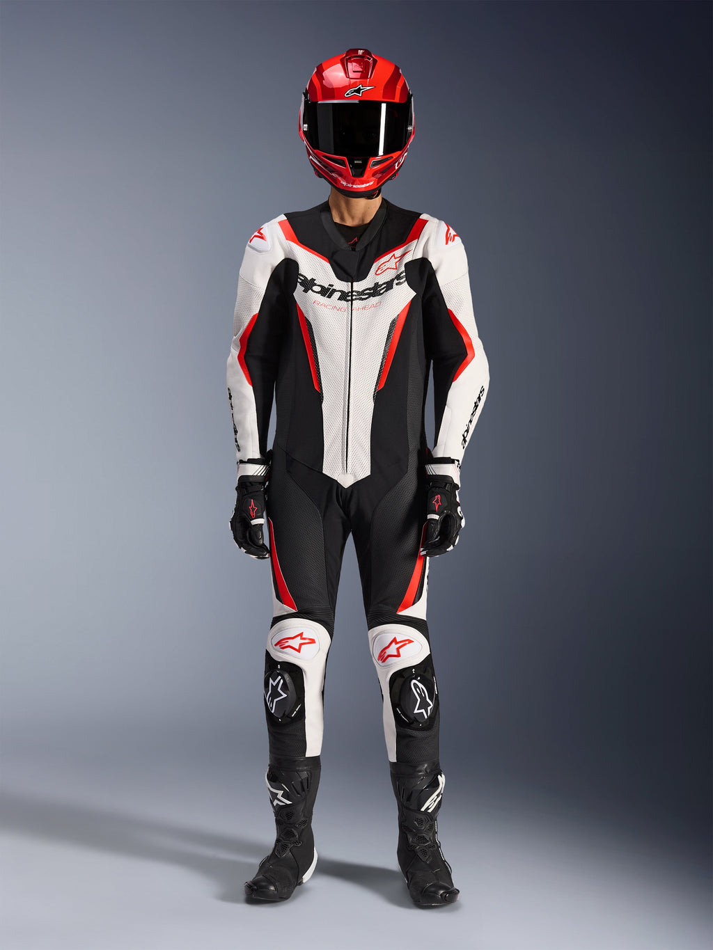 Mono de cuero Alpinestars GP Force V2 de 1 pieza, blanco, negro y rojo flúor, equipamiento completo de carreras que incluye casco rojo, guantes y botas negros, con diseño aerodinámico y logotipos de Alpinestars en el pecho y las rodillas