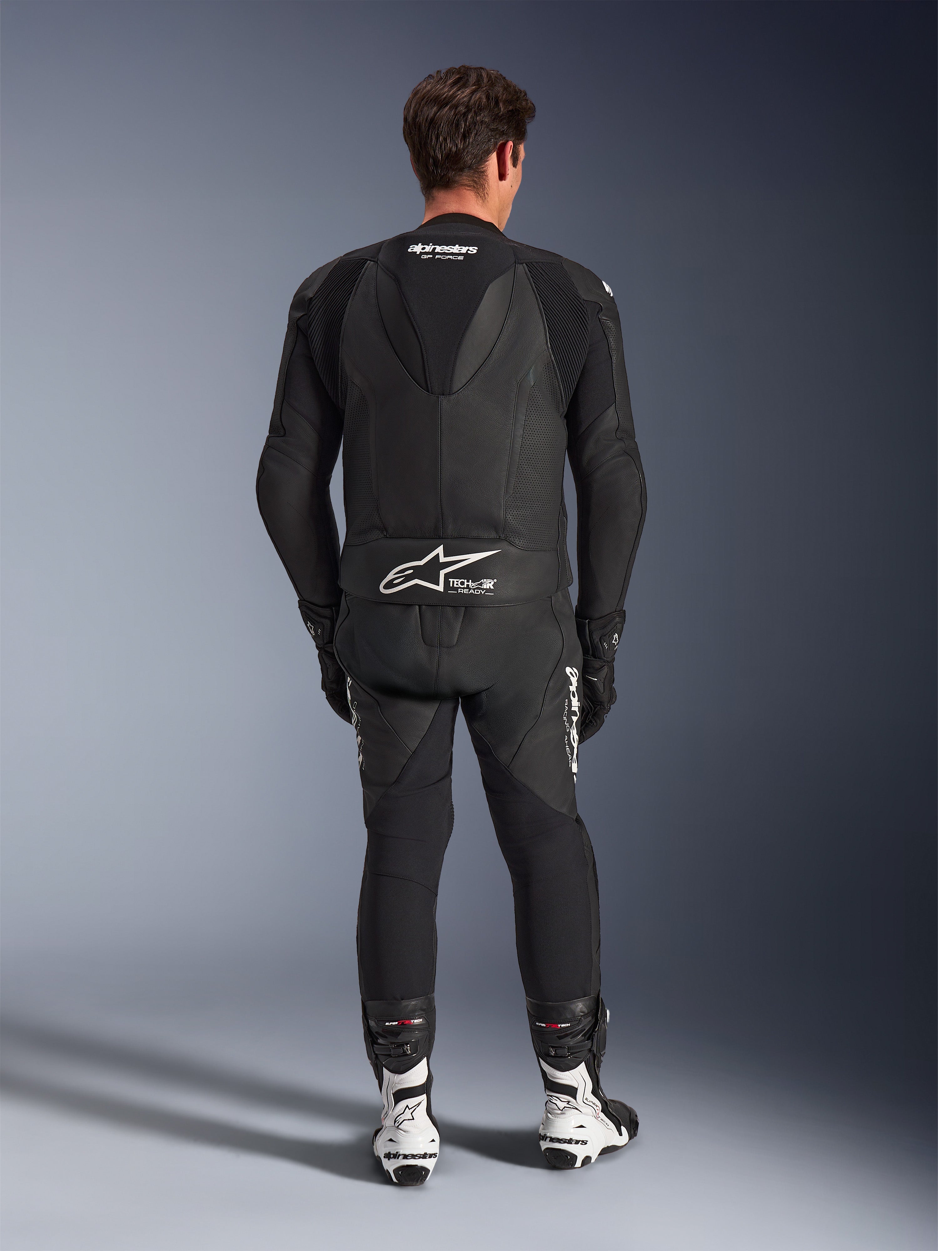Traje de cuero Alpinestars GP Force V2 de 2 piezas, negro, vista trasera que muestra la joroba aerodinámica y el logotipo Tech-Air Ready en la cintura, paneles de cuero perforado para ventilación, combinado con botas de competición blancas y negras, equipamiento profesional para carreras de motociclismo