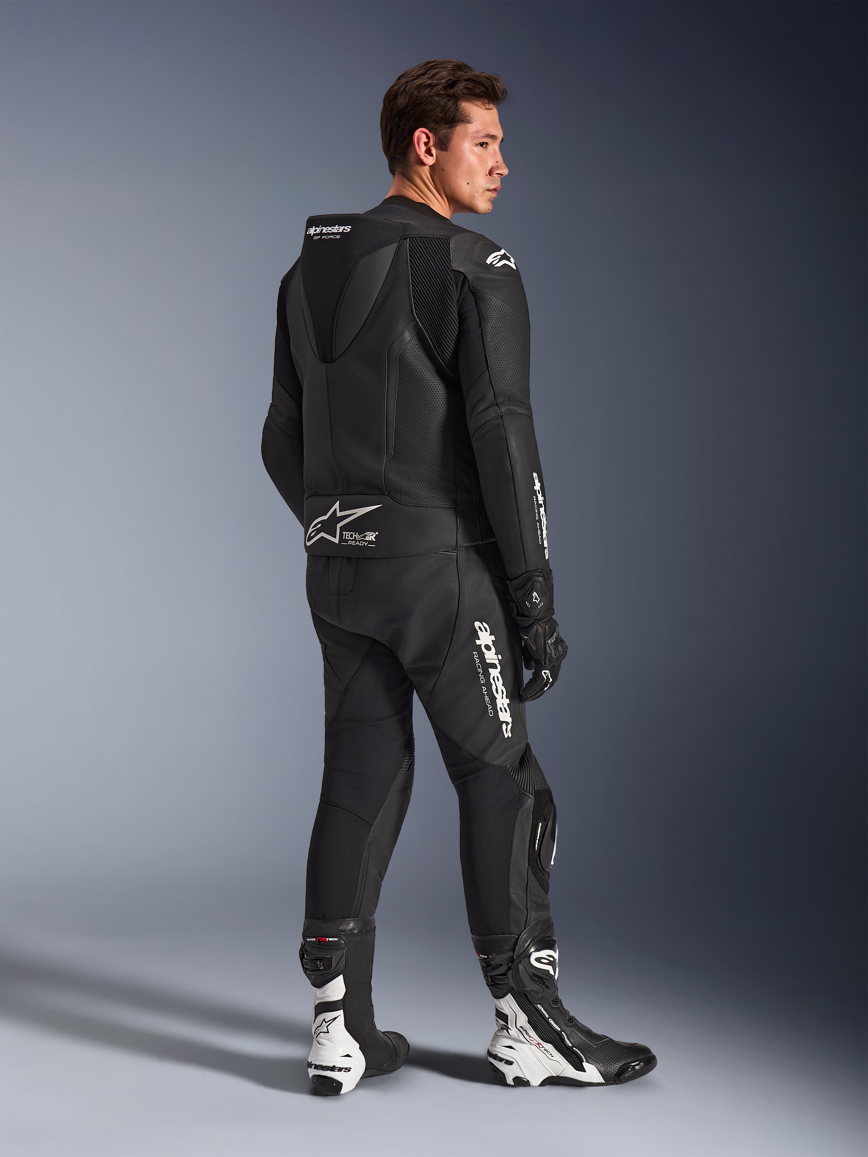 Modelo masculino con traje de cuero Alpinestars GP Force V2 de 2 piezas en negro con logotipos blancos, vista trasera de tres cuartos que muestra la joroba aerodinámica y el distintivo Tech-Air Ready, combinado con botas y guantes de carreras en blanco y negro, equipamiento profesional para motociclismo deportivo y de pista.
