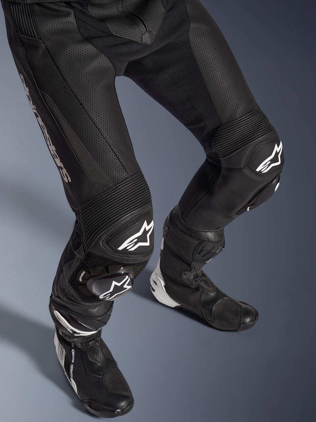 Alpinestars GP Force V2 2-Piece Leather Suit, primer plano de la parte inferior del cuerpo incluyendo pantalones y botas de carreras, negro con logotipos blancos, con construcción de cuero perforado, paneles elásticos de acordeón en las rodillas y deslizadores protectores, equipamiento profesional para conducción deportiva.