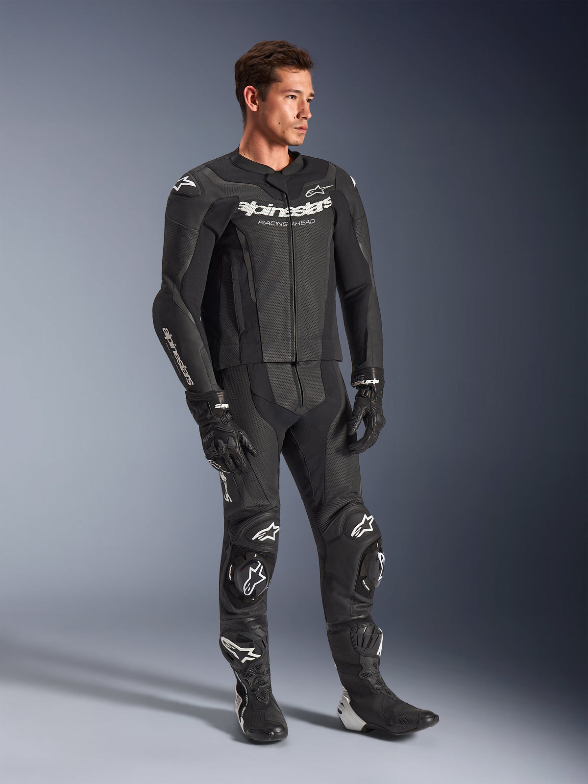 Traje de cuero de 2 piezas Alpinestars GP Force V2, negro, usado por un modelo masculino con guantes y botas de carreras negros a juego, con logotipos de Alpinestars en blanco, paneles de cuero perforado para ventilación y deslizaderas de rodilla integradas, equipo profesional de carreras de motos