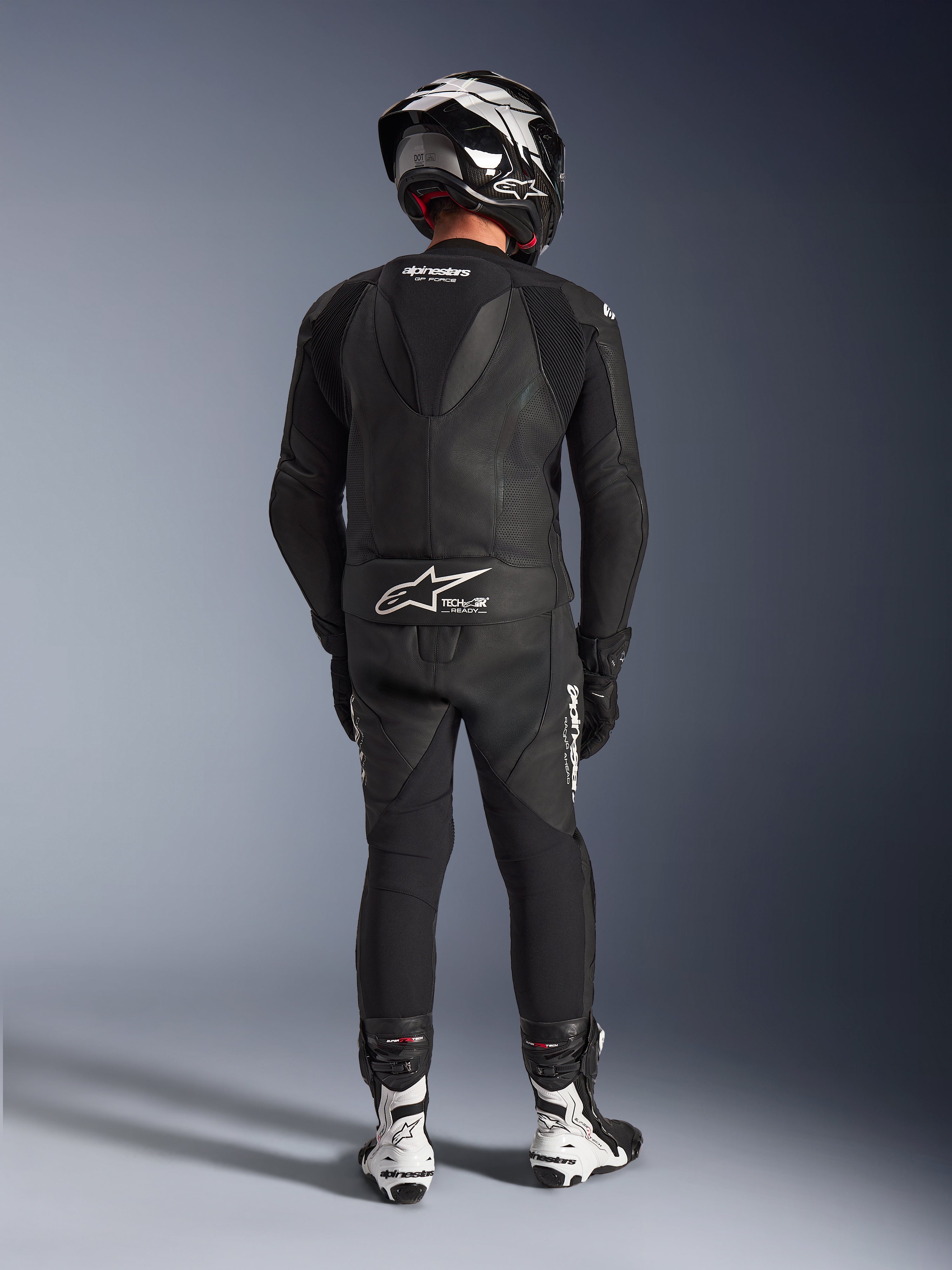 Alpinestars GP Force V2 2-Piece Leather Suit en color negro, vista trasera completa que muestra la joroba aerodinámica y la marca Tech-Air Ready, combinado con casco a juego en blanco y negro y botas de carreras blancas para un rendimiento profesional en pista