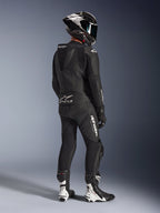 Alpinestars GP Force V2 2-Piece Leather Suit, negro, vista posterior de un piloto de motociclismo con equipamiento de competición completo, con joroba aerodinámica, logotipos blancos y cuero perforado, combinado con un casco de fibra de carbono y botas de carreras blancas y negras