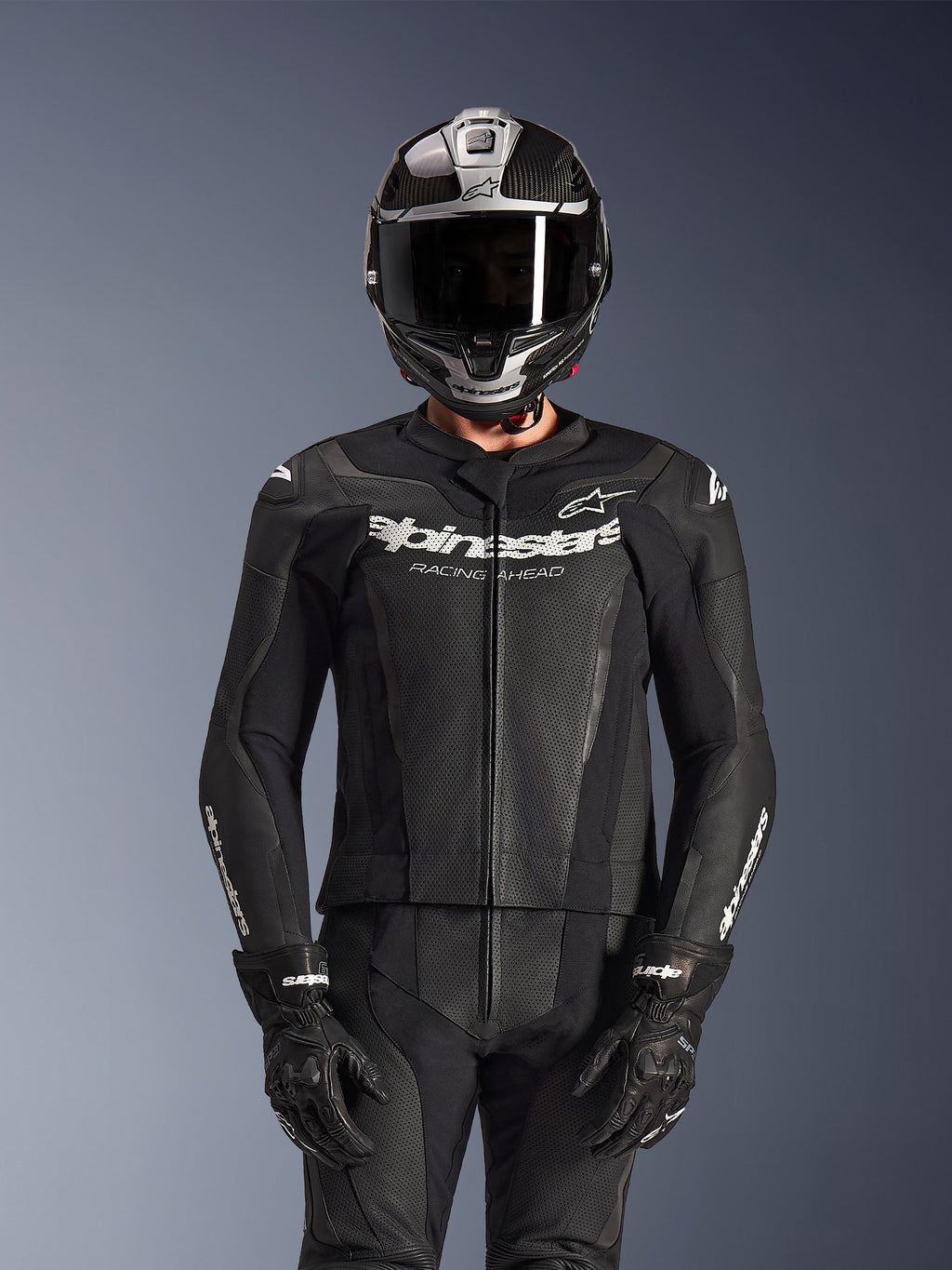 Piloto con traje de cuero de 2 piezas Alpinestars GP Force V2 en color negro, construcción de cuero perforado con logotipo 'Racing Ahead' blanco en el pecho, casco integral plateado y de fibra de carbono, guantes negros, equipamiento profesional para conducción deportiva y en pista