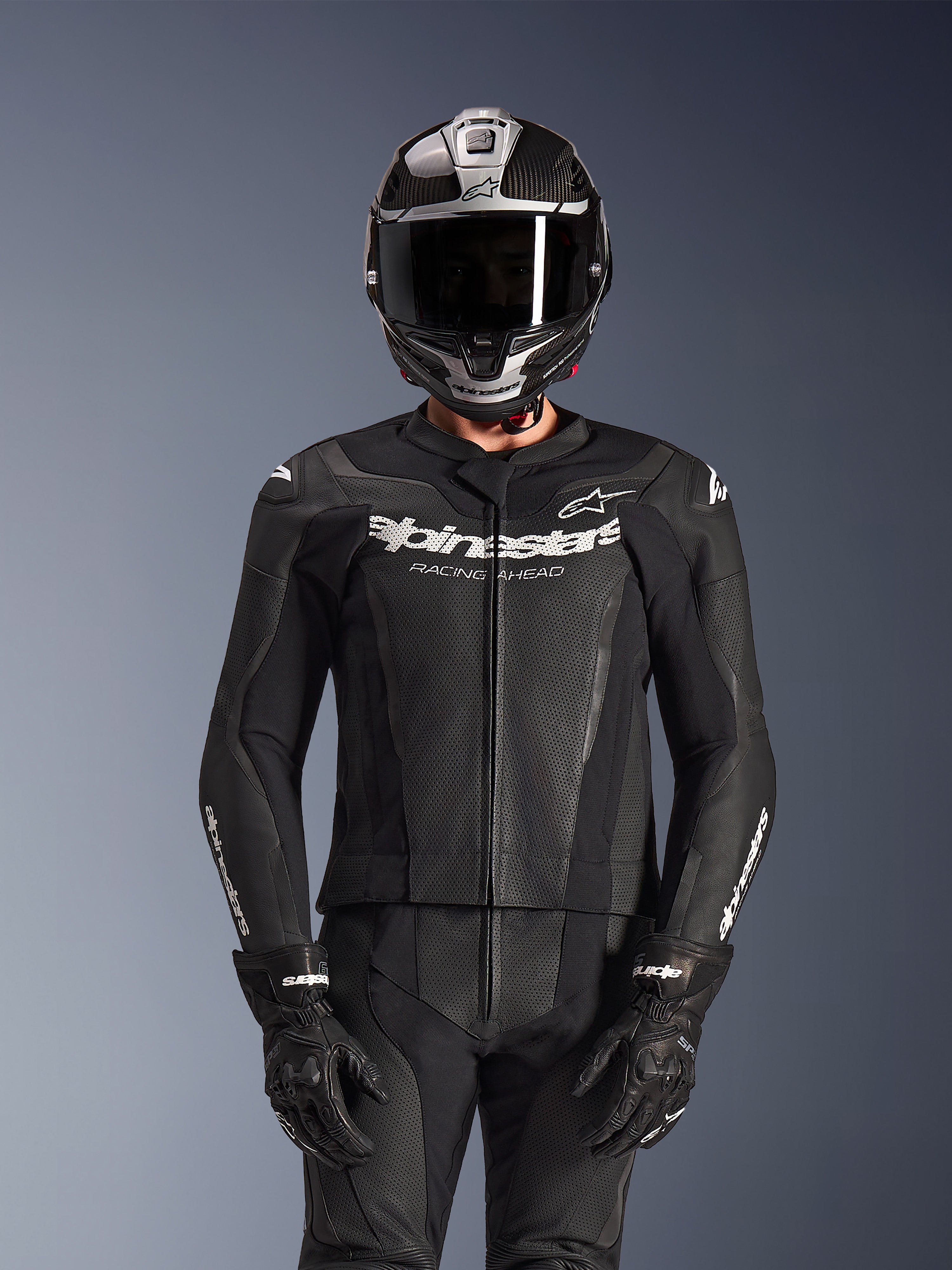 Piloto con traje de cuero de 2 piezas Alpinestars GP Force V2 en color negro, construcción de cuero perforado con logotipo 'Racing Ahead' blanco en el pecho, casco integral plateado y de fibra de carbono, guantes negros, equipamiento profesional para conducción deportiva y en pista