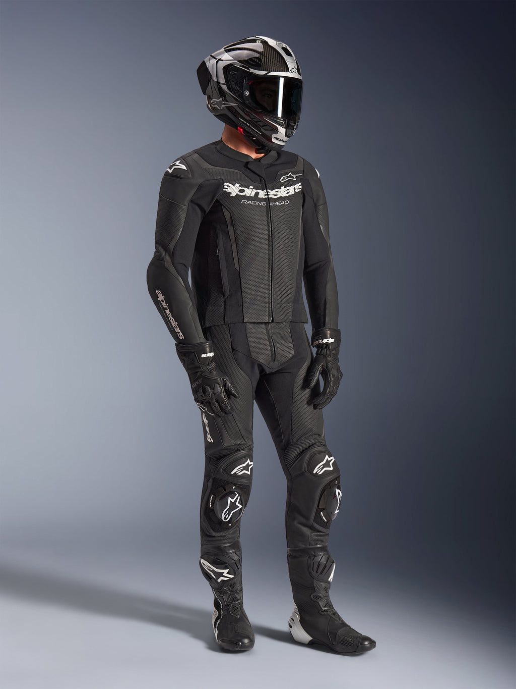 Traje de cuero de 2 piezas Alpinestars GP Force V2, traje negro de carreras de motociclismo, usado con casco integral, guantes y botas, con construcción de cuero perforado, joroba aerodinámica y logotipos blancos de Alpinestars en el pecho, brazos y rodillas.