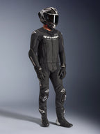 Traje de cuero de 2 piezas Alpinestars GP Force V2, traje negro de carreras de motociclismo, usado con casco integral, guantes y botas, con construcción de cuero perforado, joroba aerodinámica y logotipos blancos de Alpinestars en el pecho, brazos y rodillas.