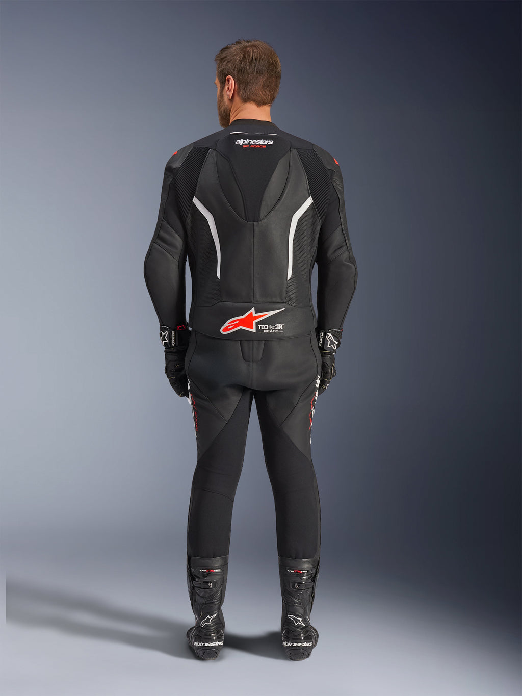 Vista trasera de un piloto con el traje de cuero de 2 piezas Alpinestars GP Force V2, negro con detalles en blanco y rojo, con logotipos prominentes de Alpinestars y Tech-Air Ready en la espalda, combinado con guantes y botas de carreras negros a juego.